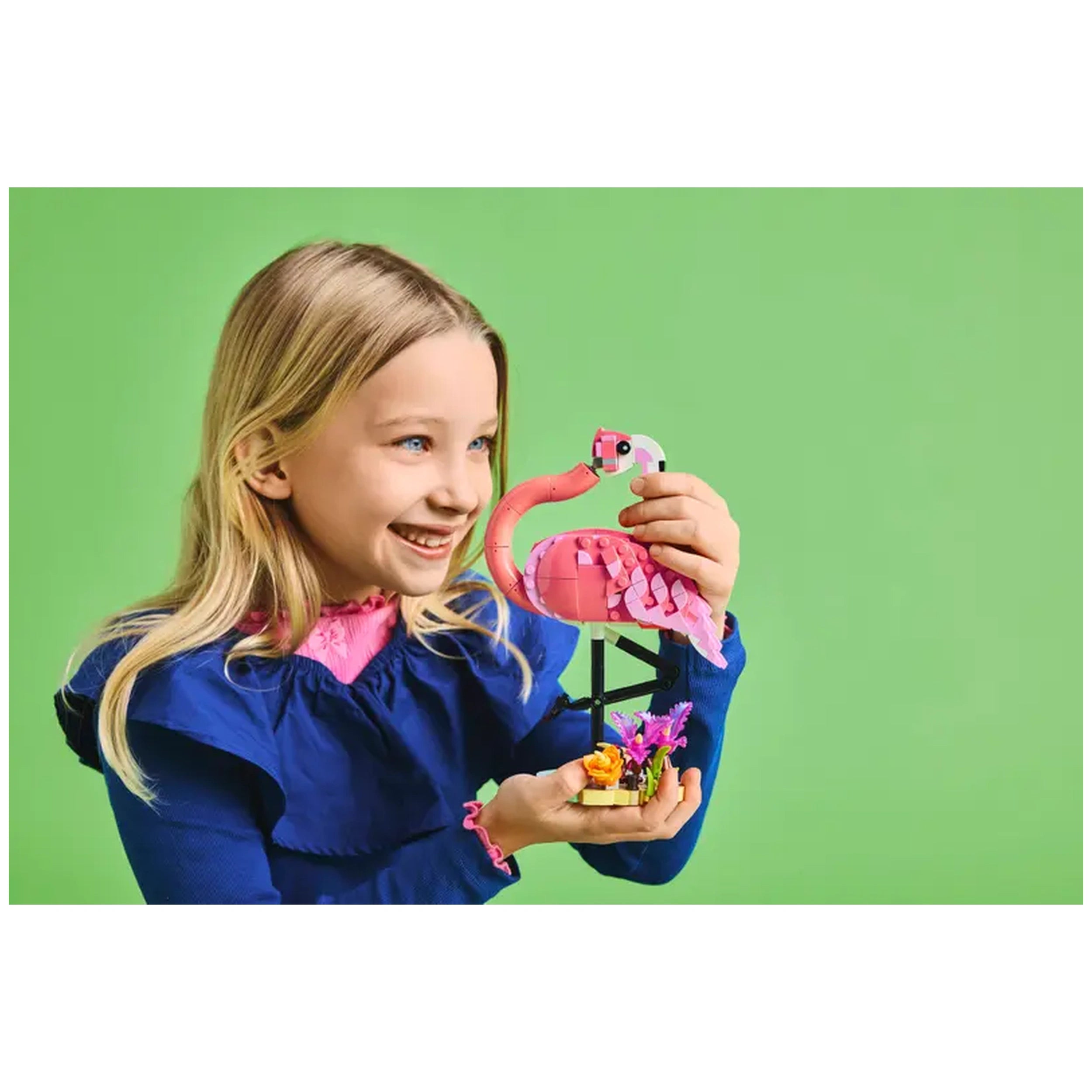 A smiling child holds a LEGO® Creator 3in1 Wild Animals pink flamingo, standing on a plant-covered base, against a bright green background.