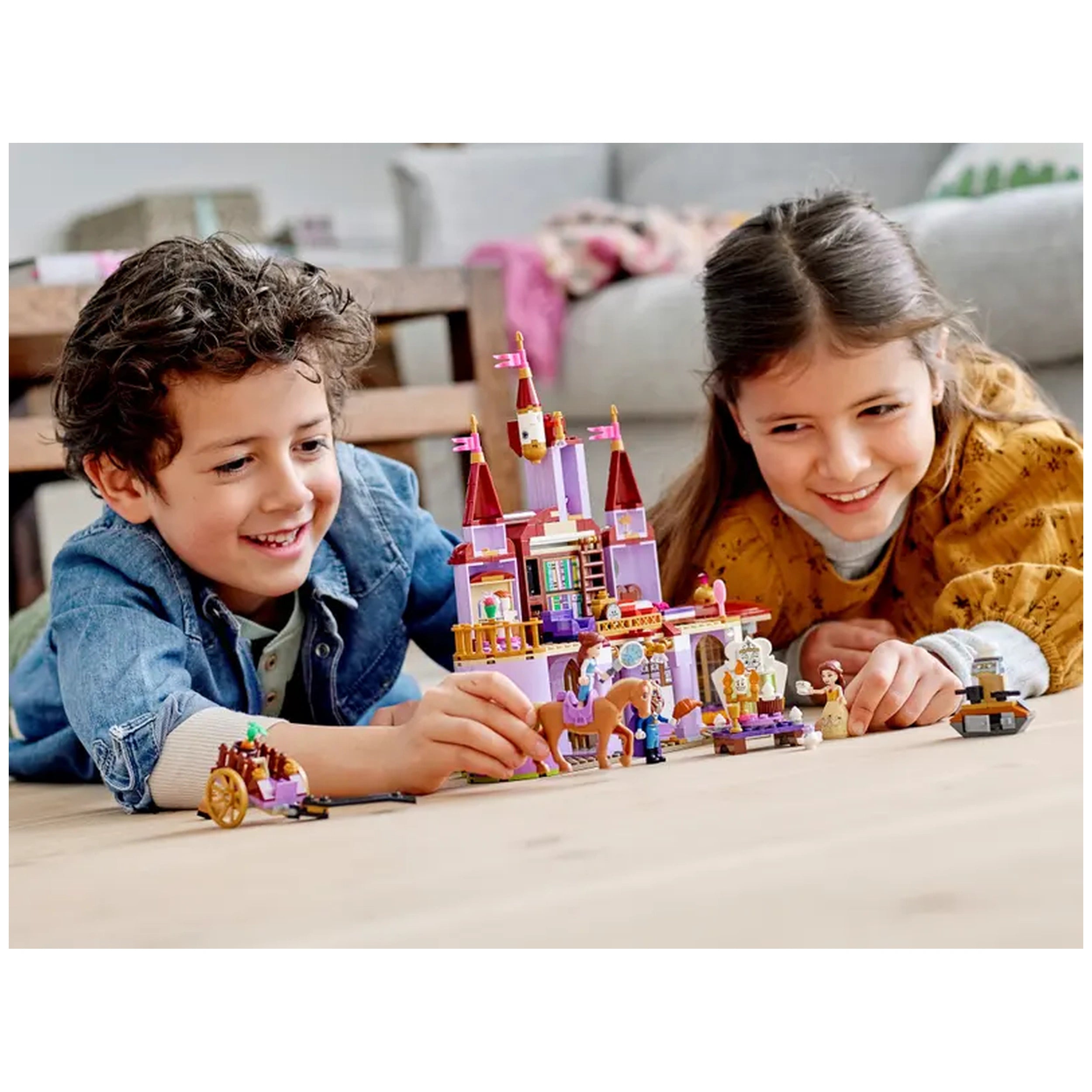Two children play on a wooden floor, focused on building the LEGO Disney Belle and the Beast’s Castle. The colorful castle features towers and flags, surrounded by LEGO figures of Belle, the Beast, and Philippe the horse. One child smiles as he adjusts the castle, while the other, wearing a patterned sweater, gestures excitedly. Soft lighting and a cozy living room setting enhance the joyful scene.