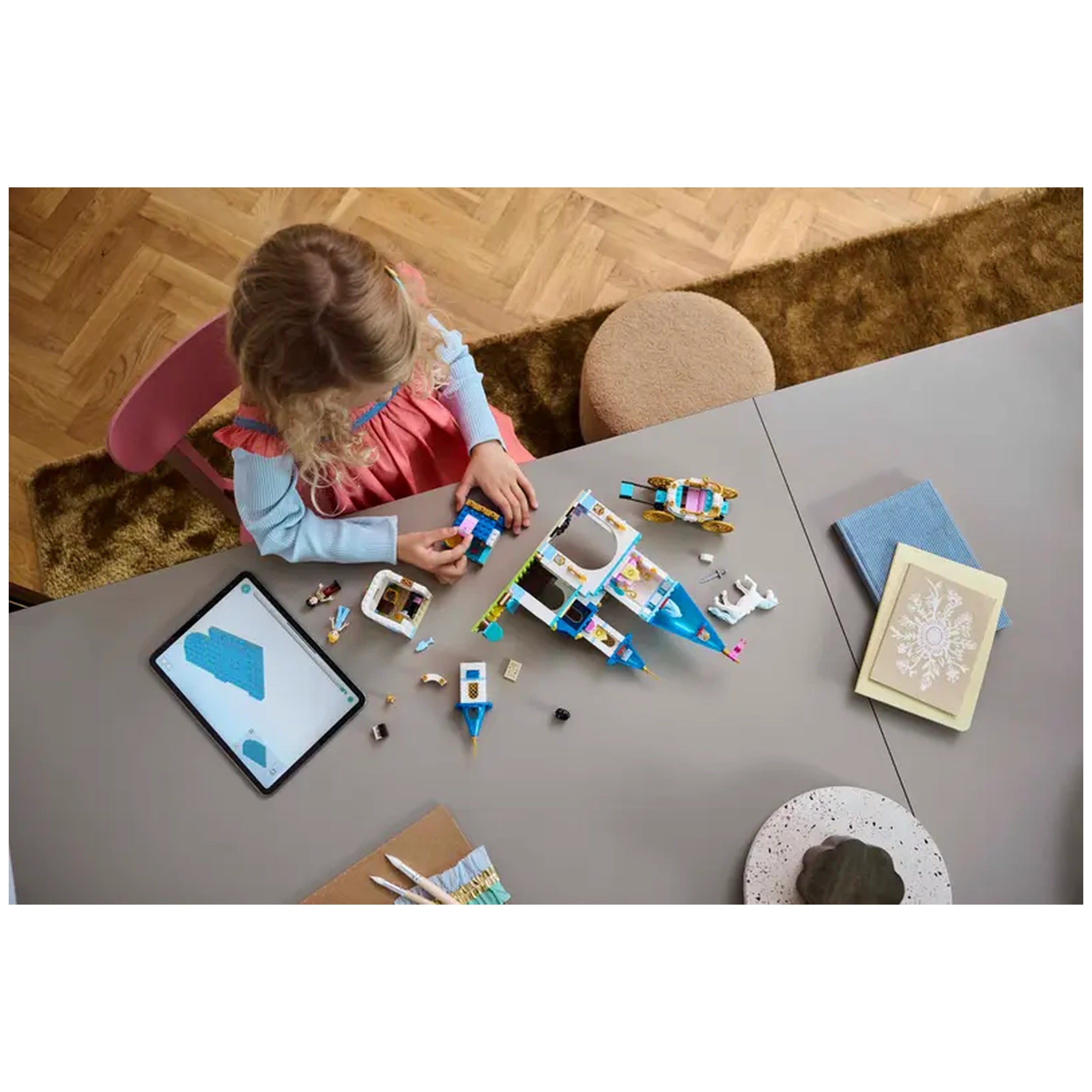A young child with curly hair builds Lego pieces on a gray table, surrounded by colorful bricks and small figures. An iPad displays building instructions, while craft supplies and a decorative book lie nearby.