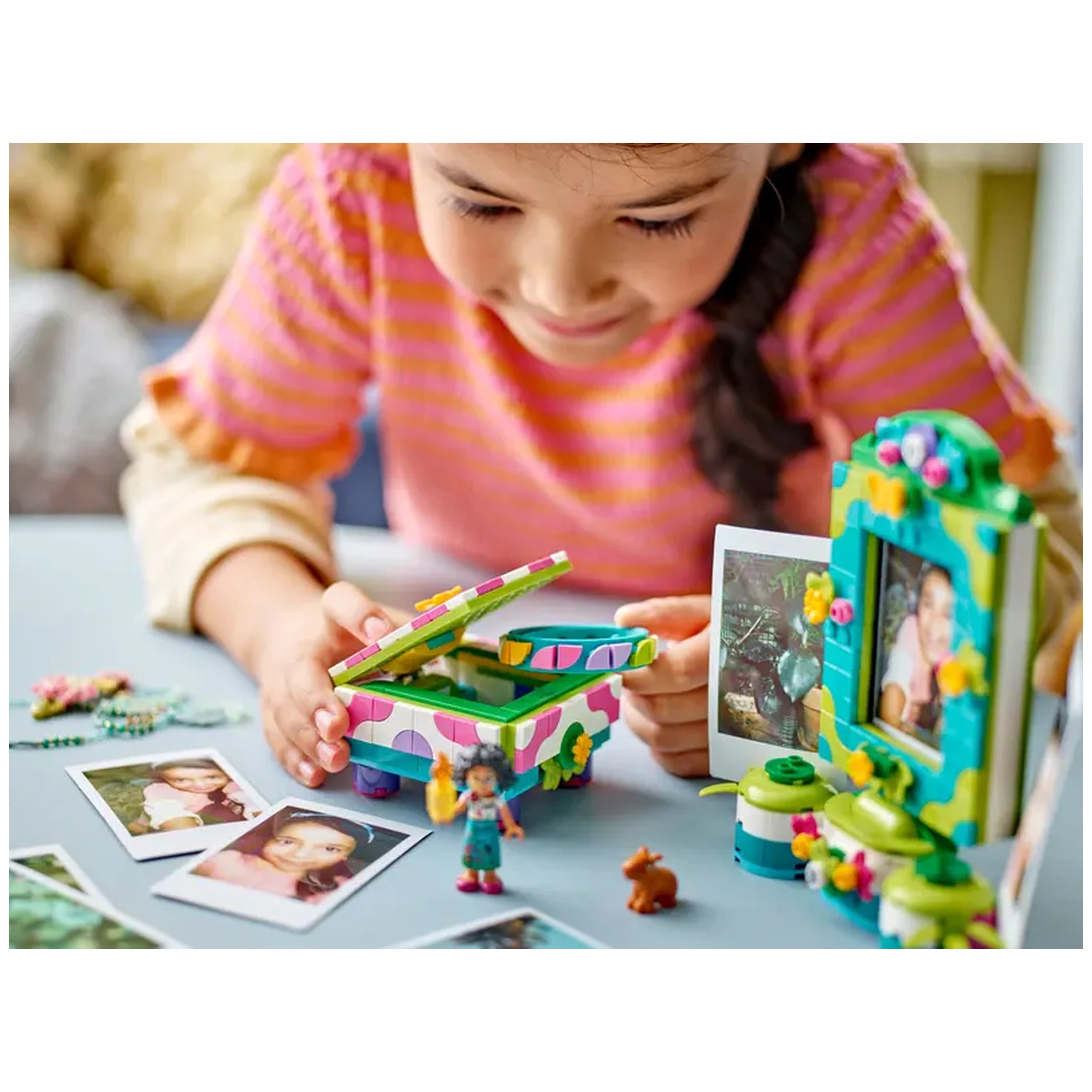A cheerful girl in a striped pink shirt opens a colorful LEGO jewelry box, surrounded by photos and a mini-doll figure.
