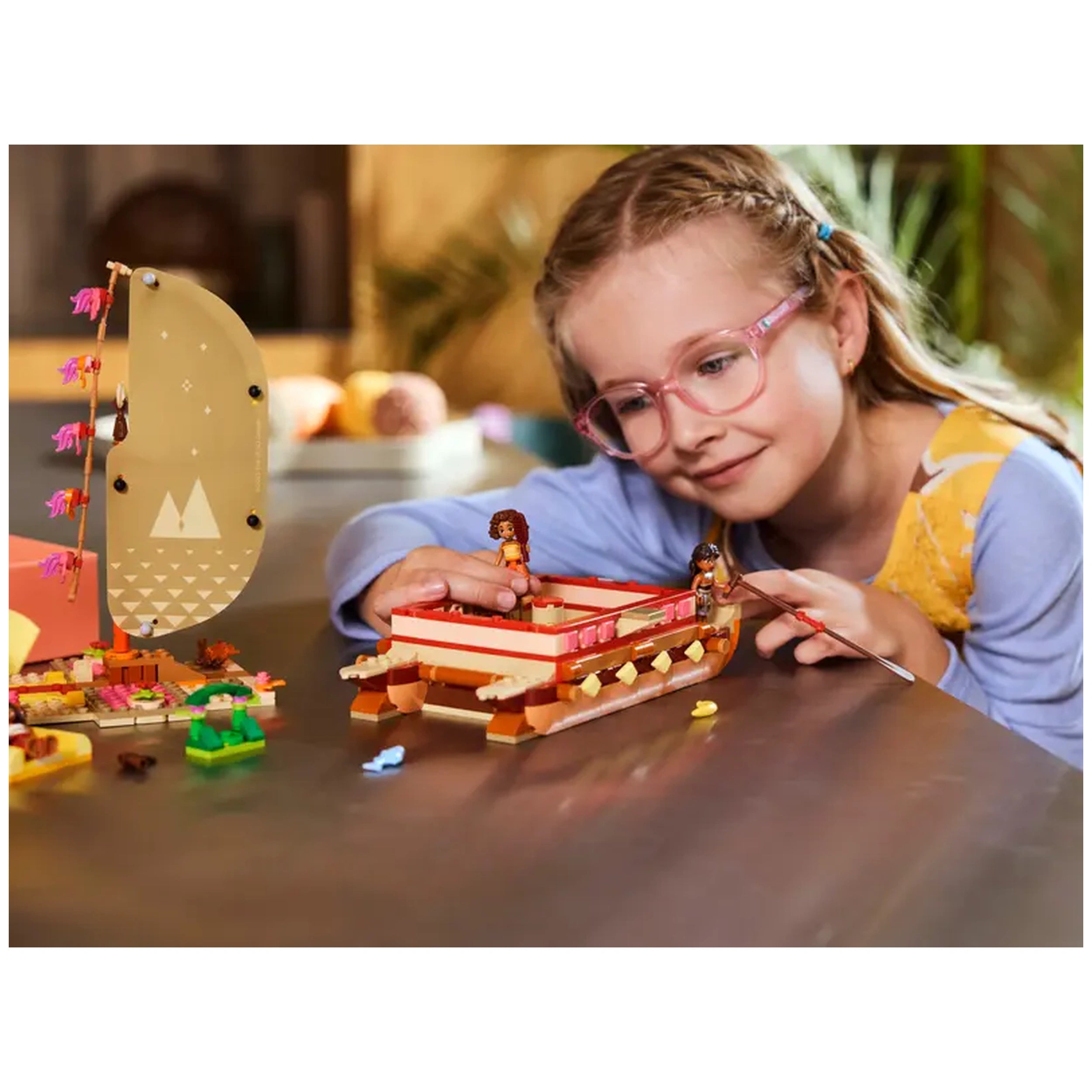 A child with glasses joyfully plays with the LEGO® Disney Moana's Adventure Canoe set, featuring a double-hulled canoe and colorful characters.