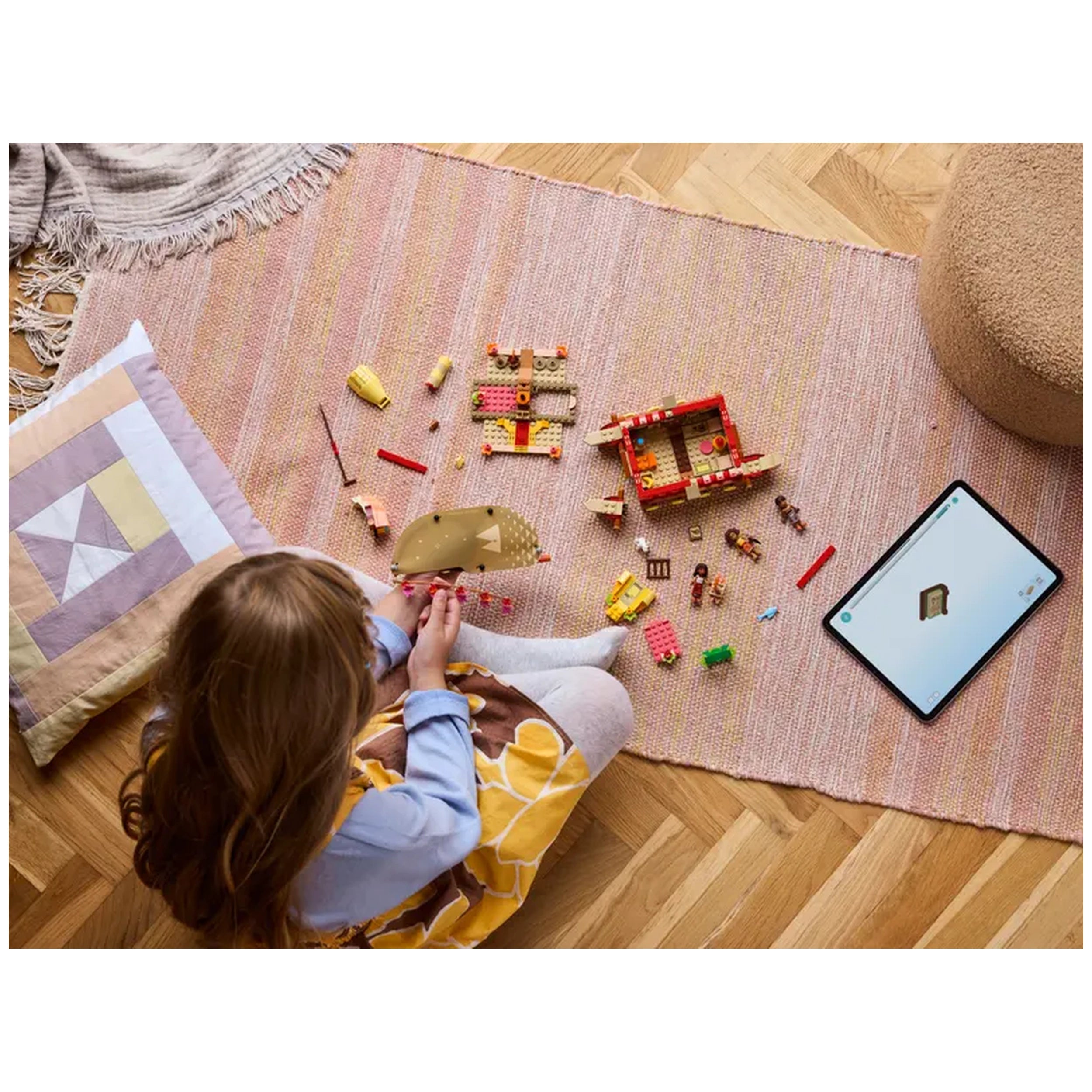 A child sits on a pink rug, assembling LEGO® pieces to create Moana's Adventure Canoe. Colorful bricks are scattered around, alongside a tablet displaying the building model. A cozy pillow sits nearby.