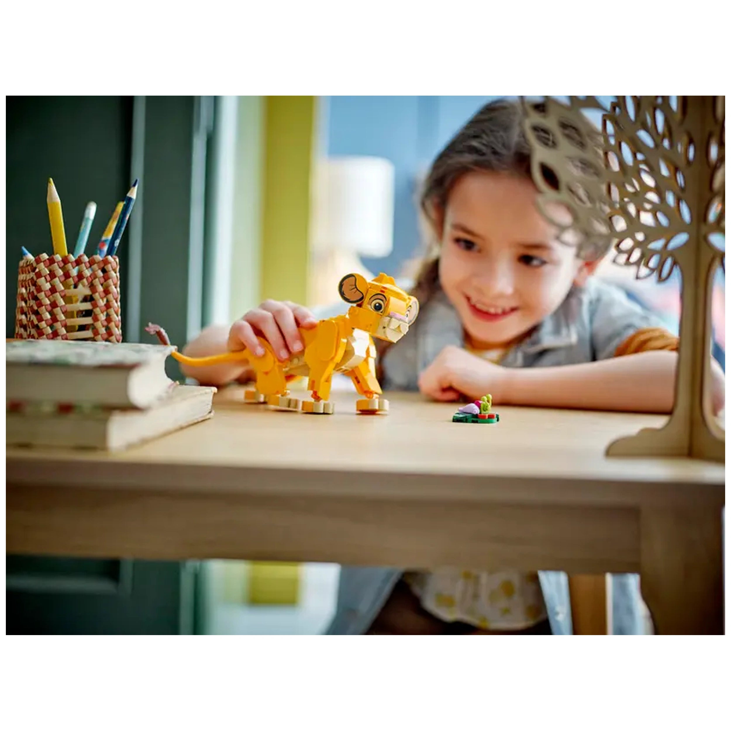 Child smiling while playing with a LEGO® Simba figure on a wooden table, alongside a small build and a pencil holder with colorful pencils.