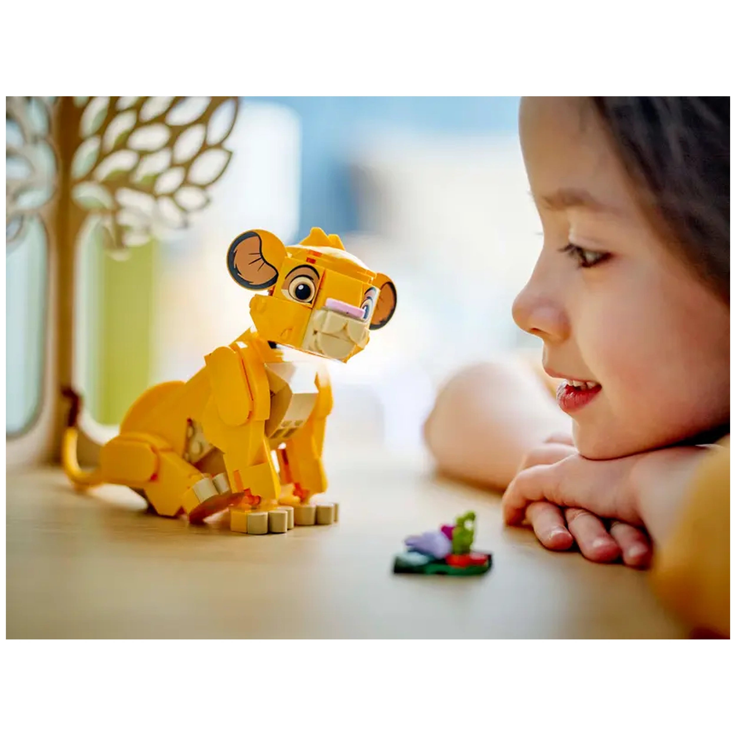 A young girl smiles while leaning on a table, gazing at a detailed yellow LEGO® Simba figure, surrounded by colorful small builds representing his food.