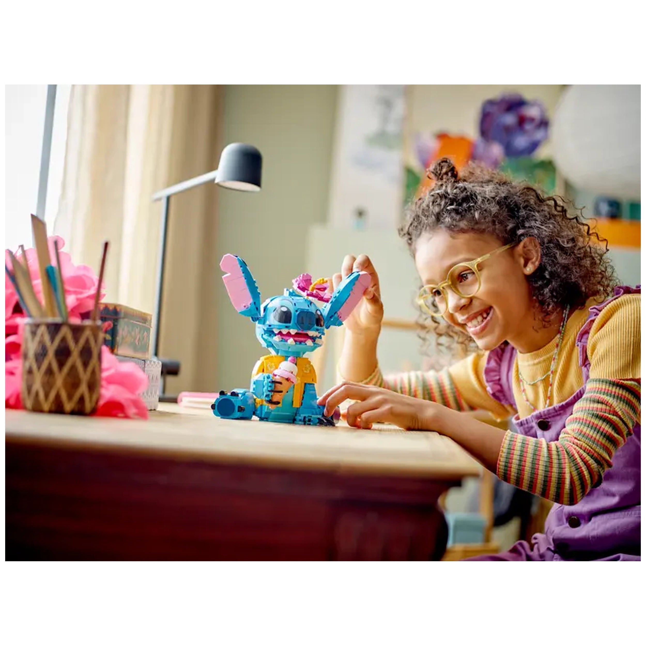 A smiling girl in stylish glasses engages with a colorful LEGO® Stitch model on a wooden table, surrounded by craft supplies.