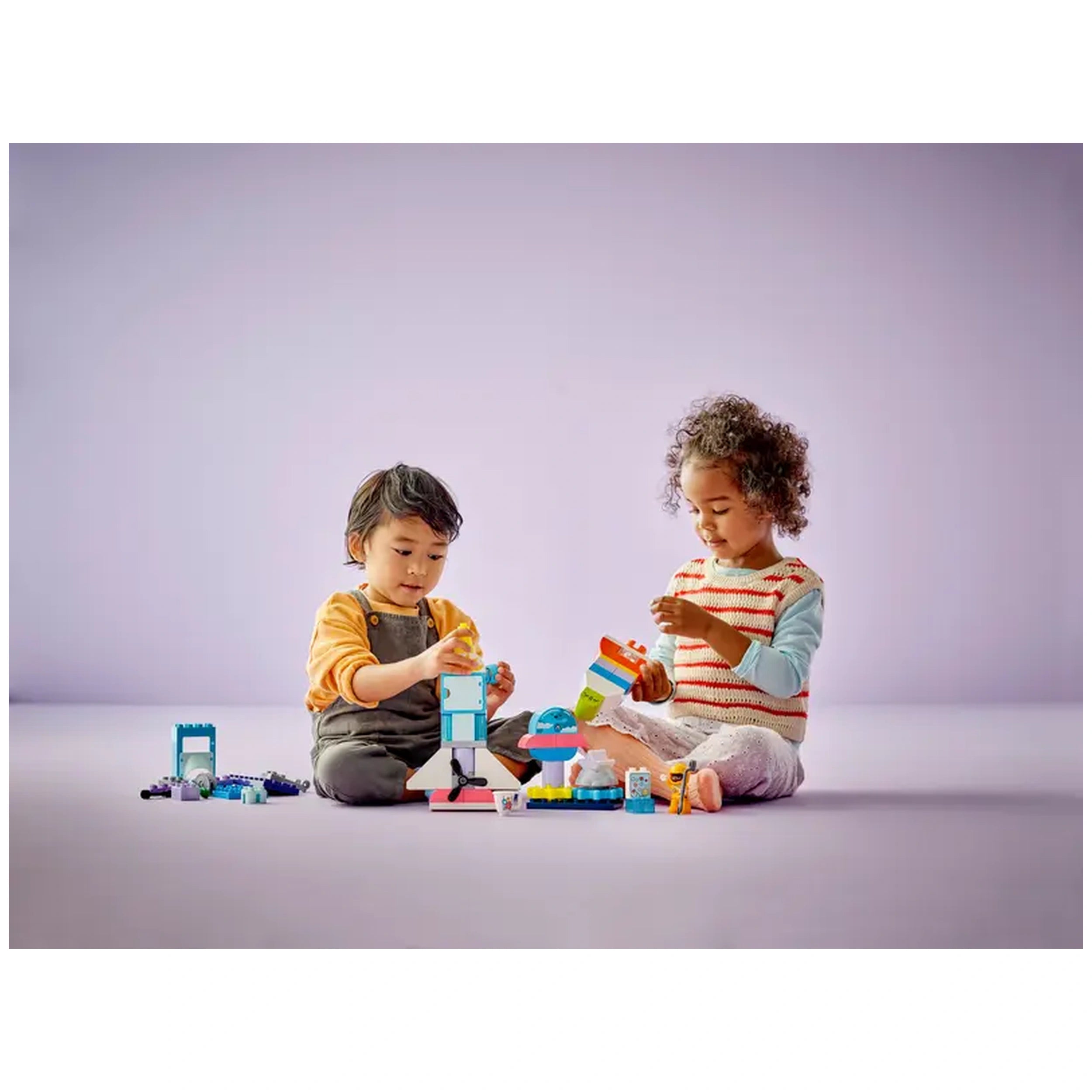 Two children play with colorful LEGO® DUPLO® blocks on a soft surface, assembling a rocket ship and a friendly alien.