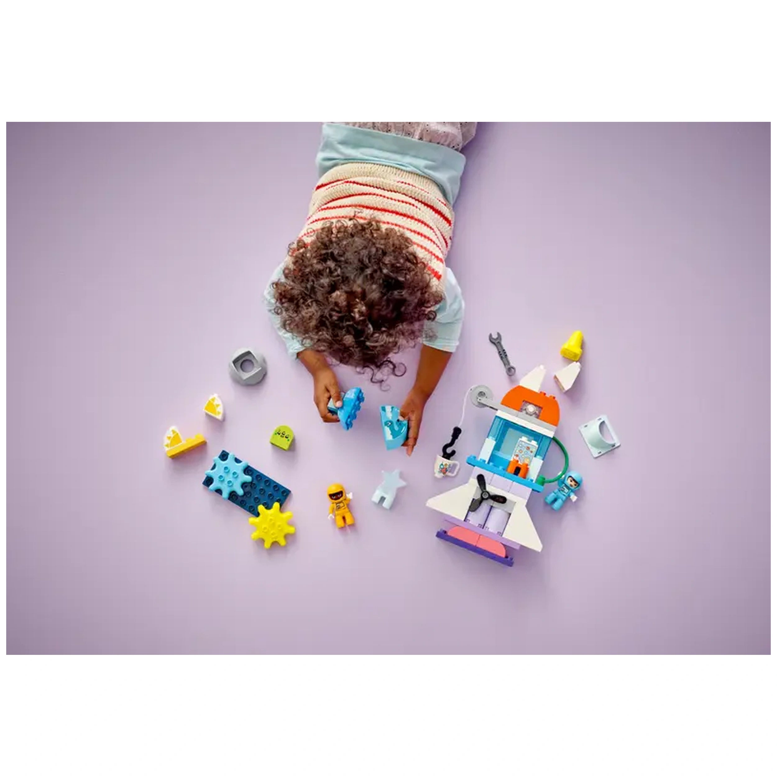 A child with curly hair kneels on a purple surface, focused on assembling colorful LEGO® DUPLO® pieces, including a rocket ship and accessories. Surrounding the child are various building blocks, astronaut figures, and playful shapes, inviting imaginative space adventures.