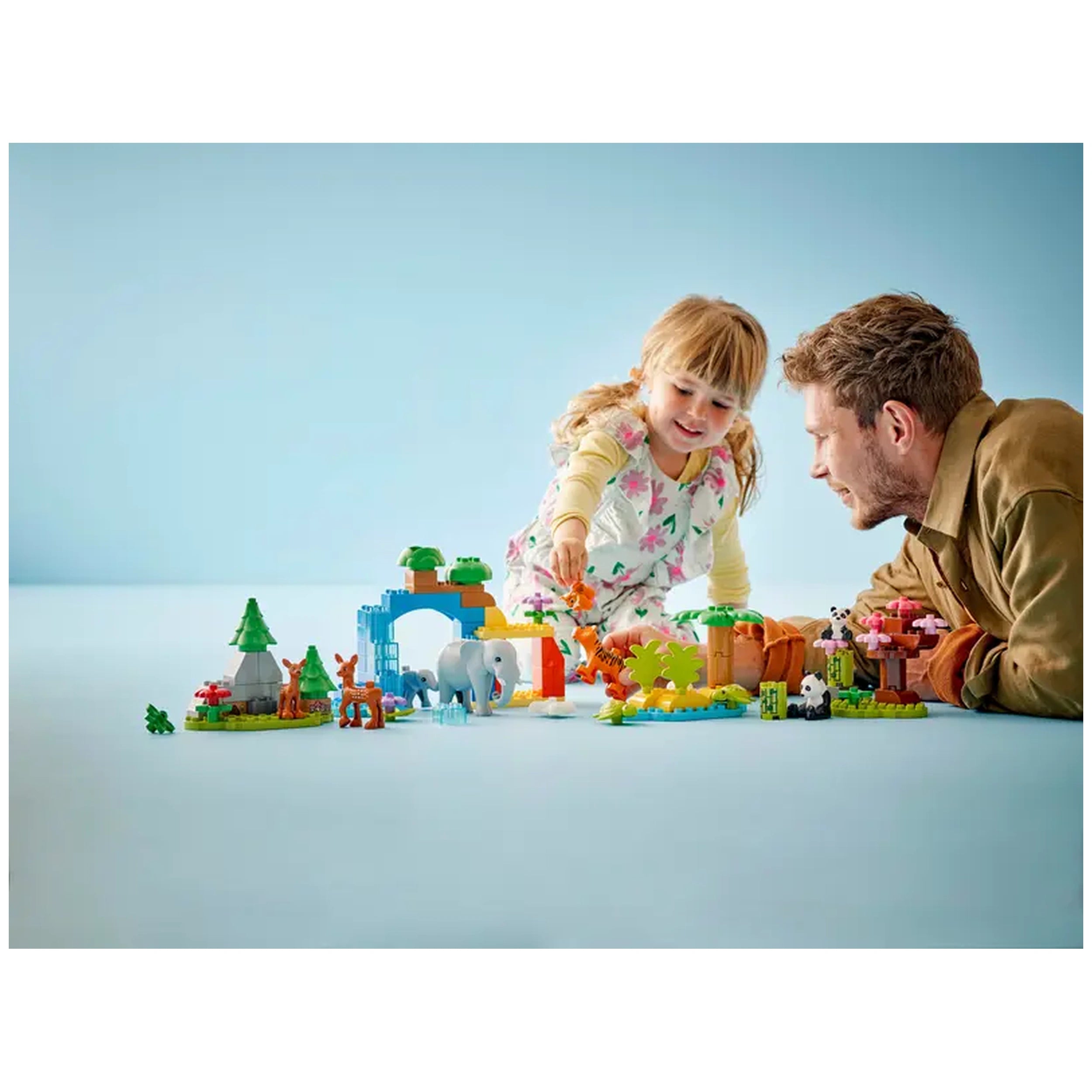 A young girl and a man joyfully play together with a colorful LEGO® DUPLO® Wild Animal Families set, featuring various animal figures and a vibrant landscape of trees and buildings.