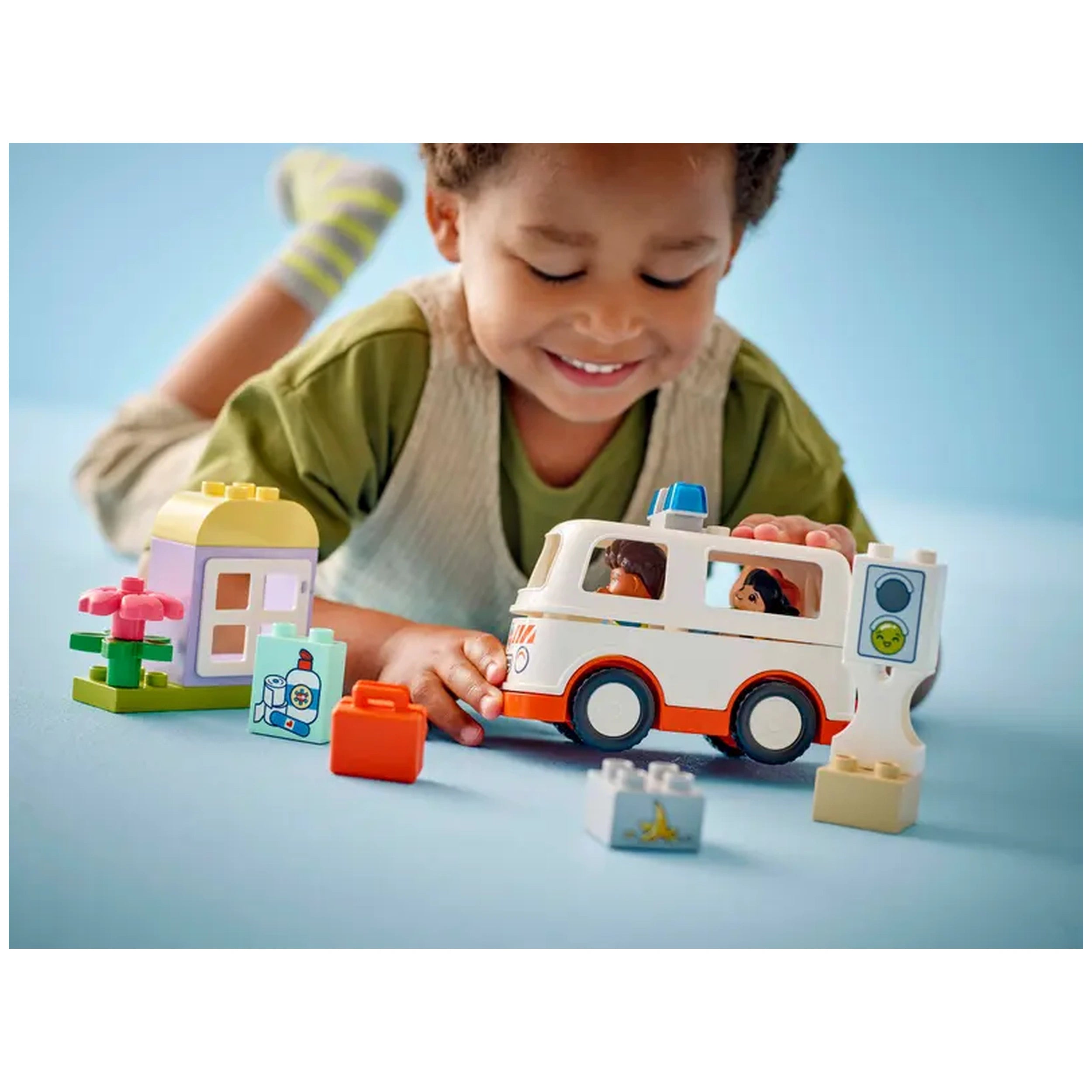 A young child smiles while playing with the LEGO® DUPLO® Ambulance & Driver set on a light blue surface. The scene features an ambulance, a flower, a small building, and various accessories, encouraging imaginative play.