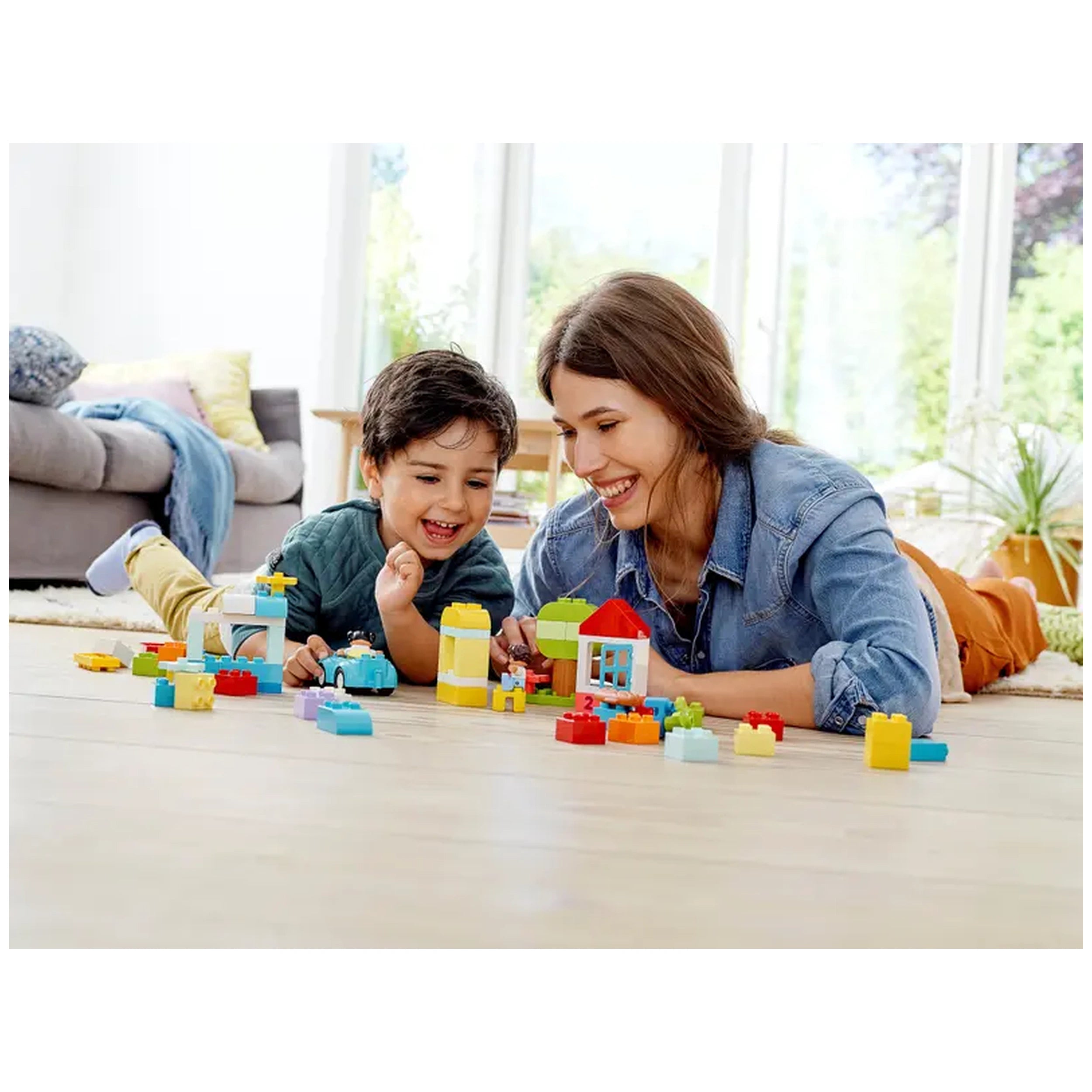 A joyful boy and woman play together on the floor, surrounded by colorful LEGO® DUPLO® pieces, including houses and cars.