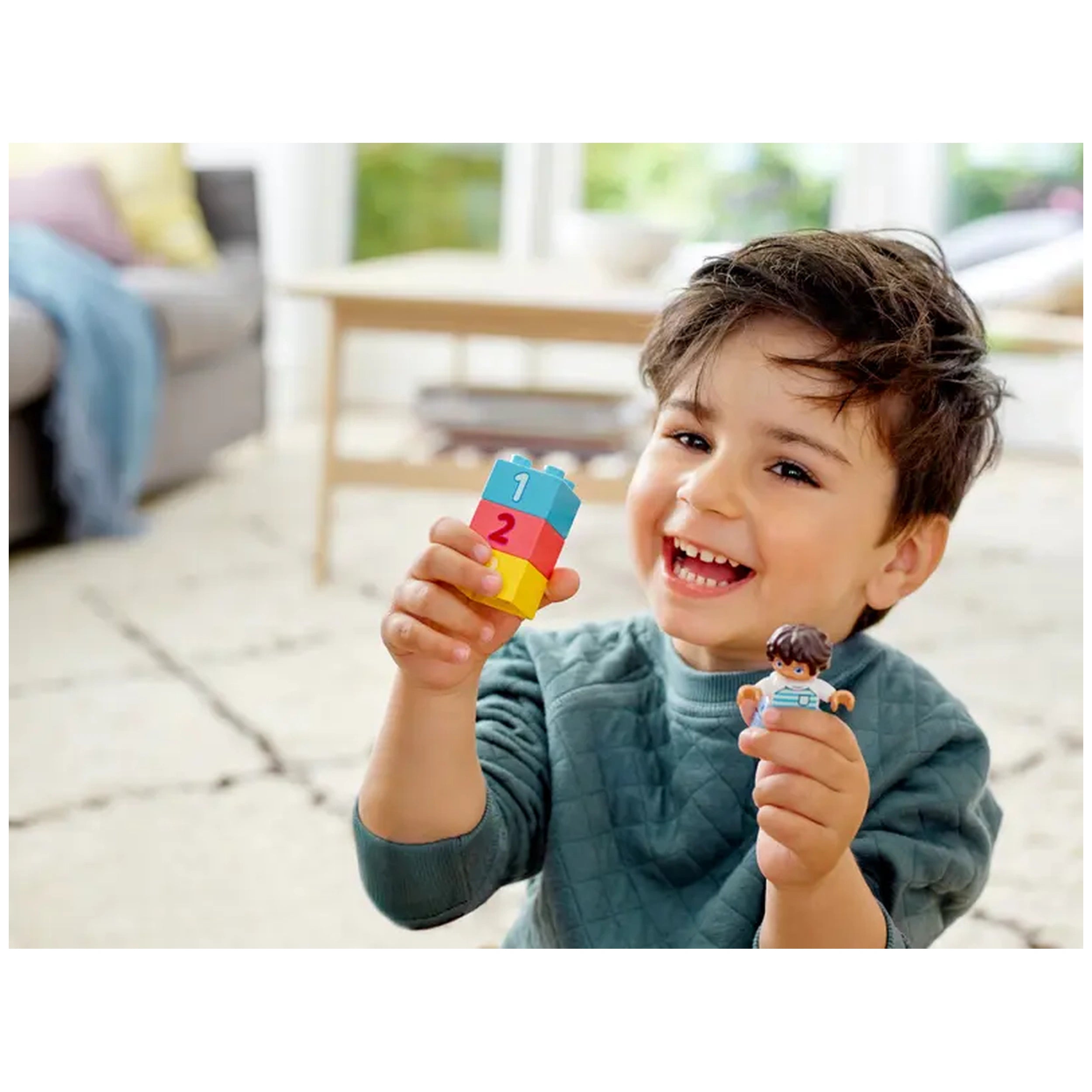 A smiling toddler holds a colorful LEGO® DUPLO® brick showing numbers '1' and '2' in one hand and a DUPLO figure in the other, sitting on a light-colored floor in a bright, cozy room.