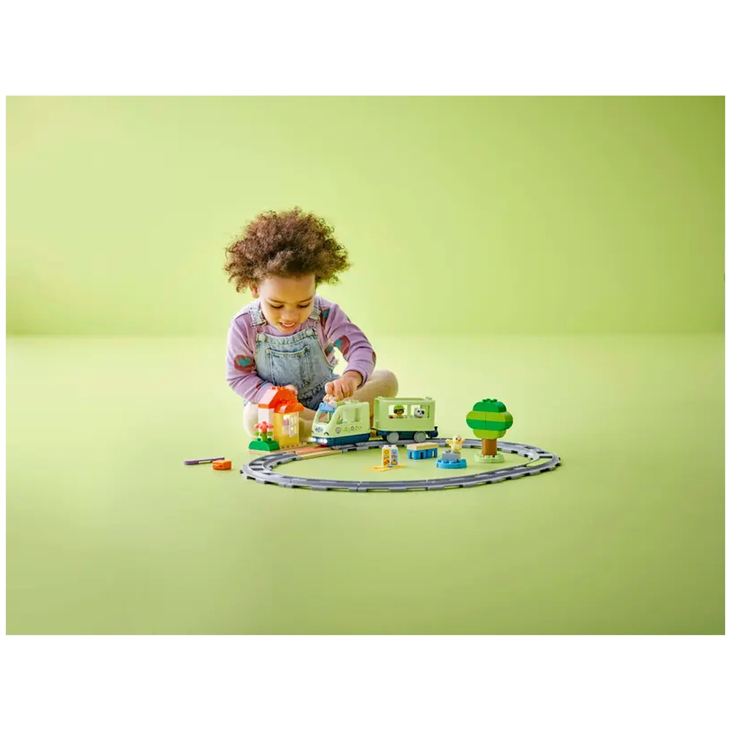A toddler with curly hair sits on a light green floor, assembling the LEGO® DUPLO® Interactive Adventure Train set, which includes a train, buildings, and a tree.