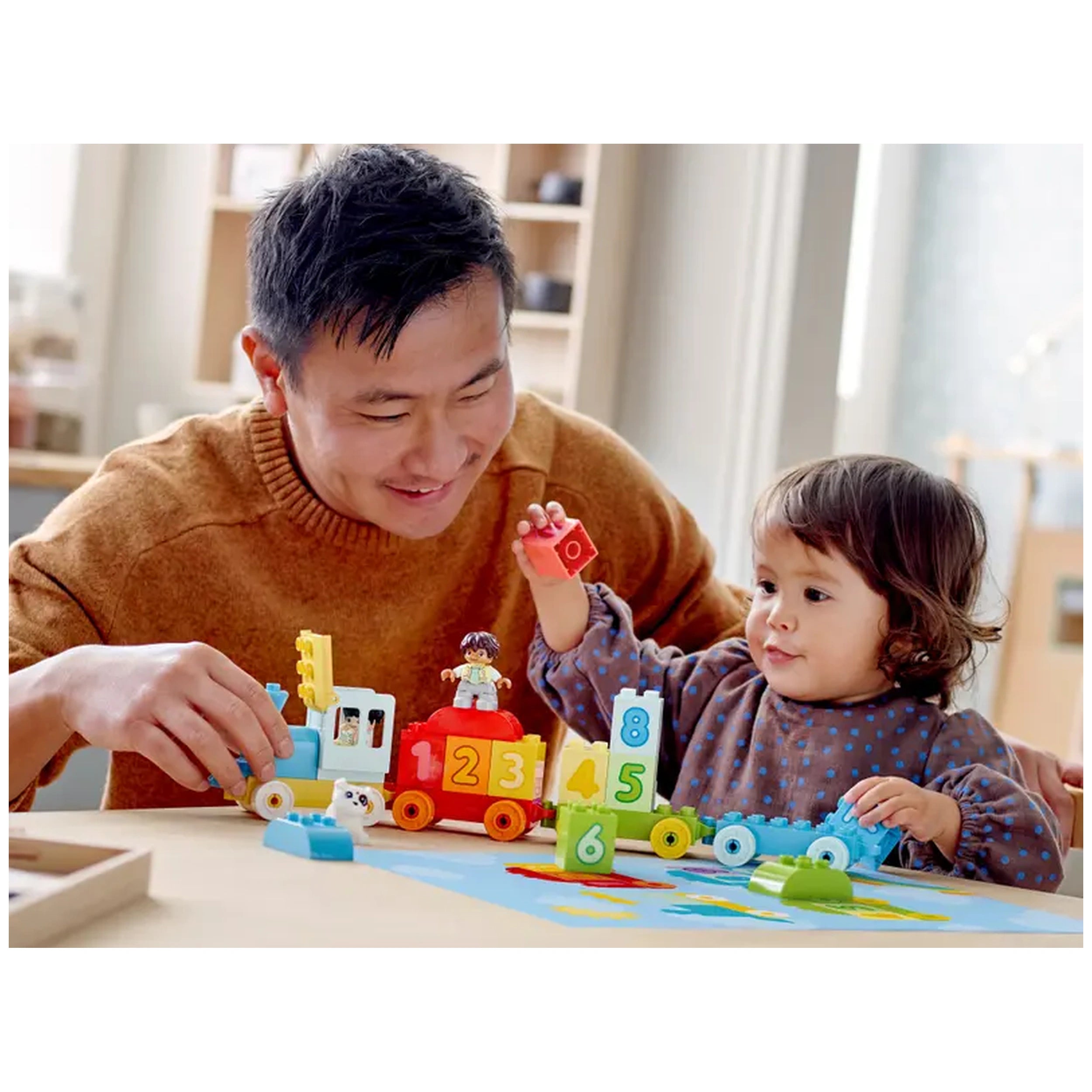 A smiling father and child play together with the LEGO® DUPLO® Number Train set. The colorful train features number bricks, while the child holds a red block. The scene is bright and inviting with various bricks scattered on a light table.