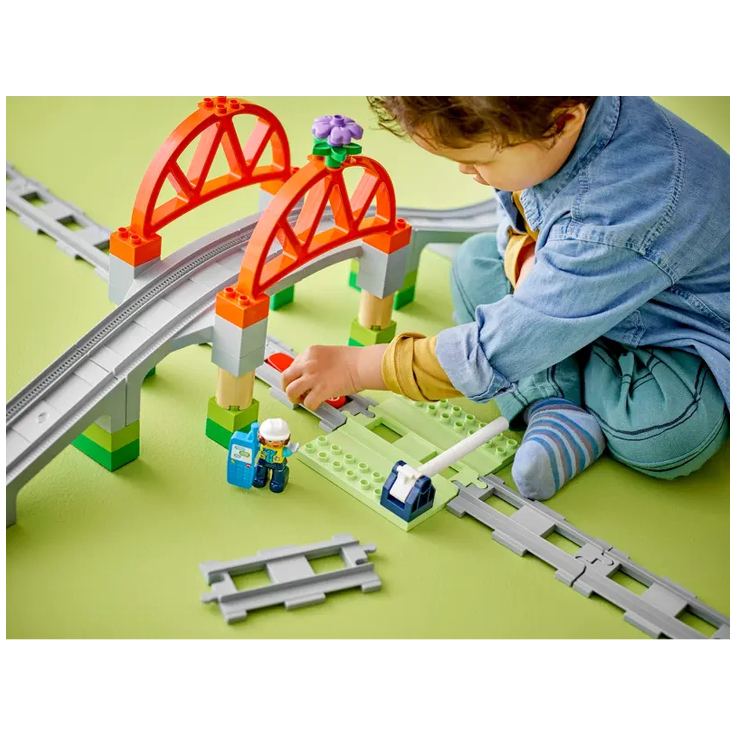 A child in a denim shirt plays with a colorful LEGO® DUPLO® Train Bridge and Tracks set, featuring a red arch bridge and a train barrier. The scene includes train tracks and small figures, set against a green background.