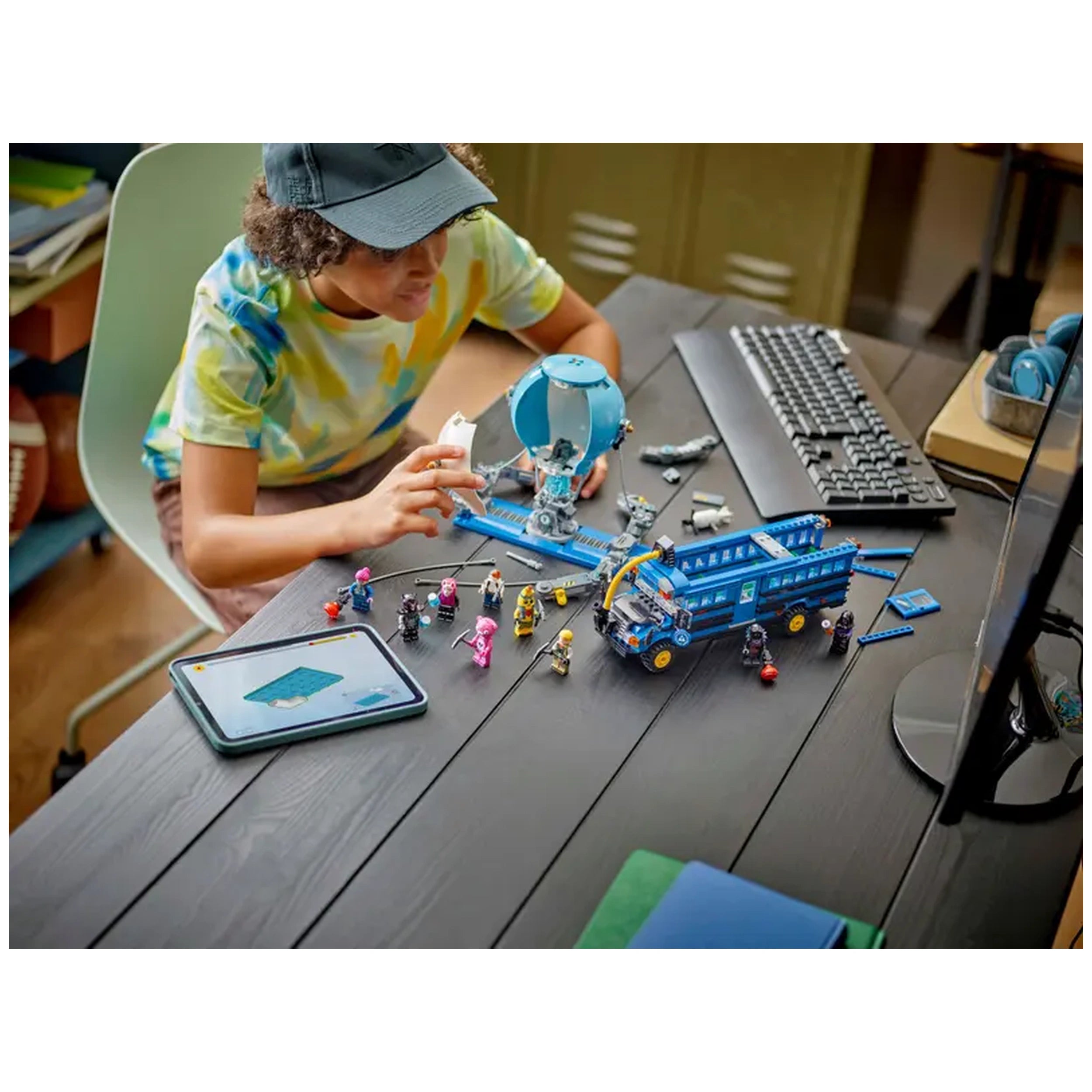 A child in a colorful shirt uses a LEGO® Fortnite Battle Bus set on a dark wooden table, surrounded by mini-figures and a tablet providing interactive building instructions. A computer keyboard and monitor are in the background.
