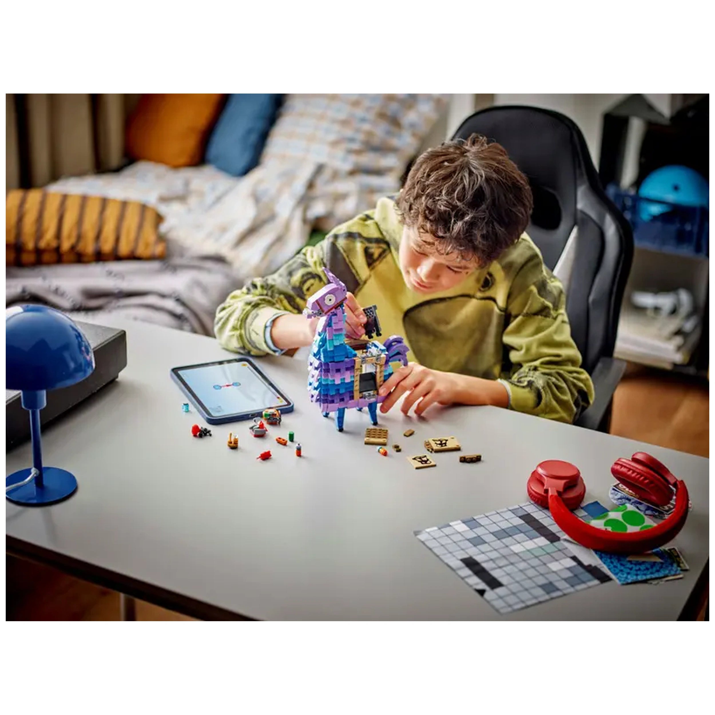 A young boy in a green hoodie builds the LEGO Fortnite Supply Llama at a desk, surrounded by colorful game accessories and a tablet.
