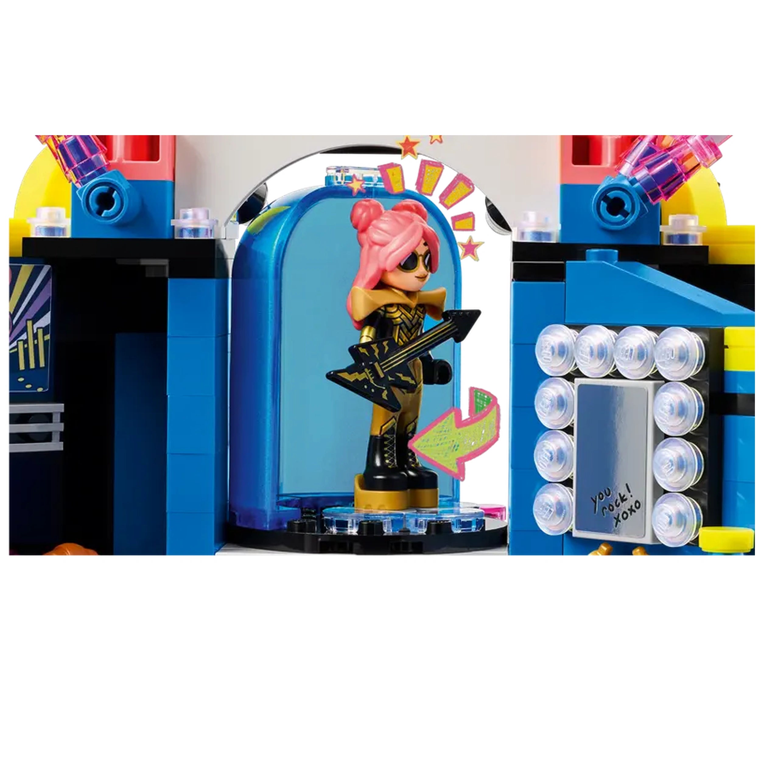 Mini-doll with pink hair in a black and gold outfit stands confidently on a stage, holding an electric guitar, surrounded by colorful set details, including lights and a mirror.