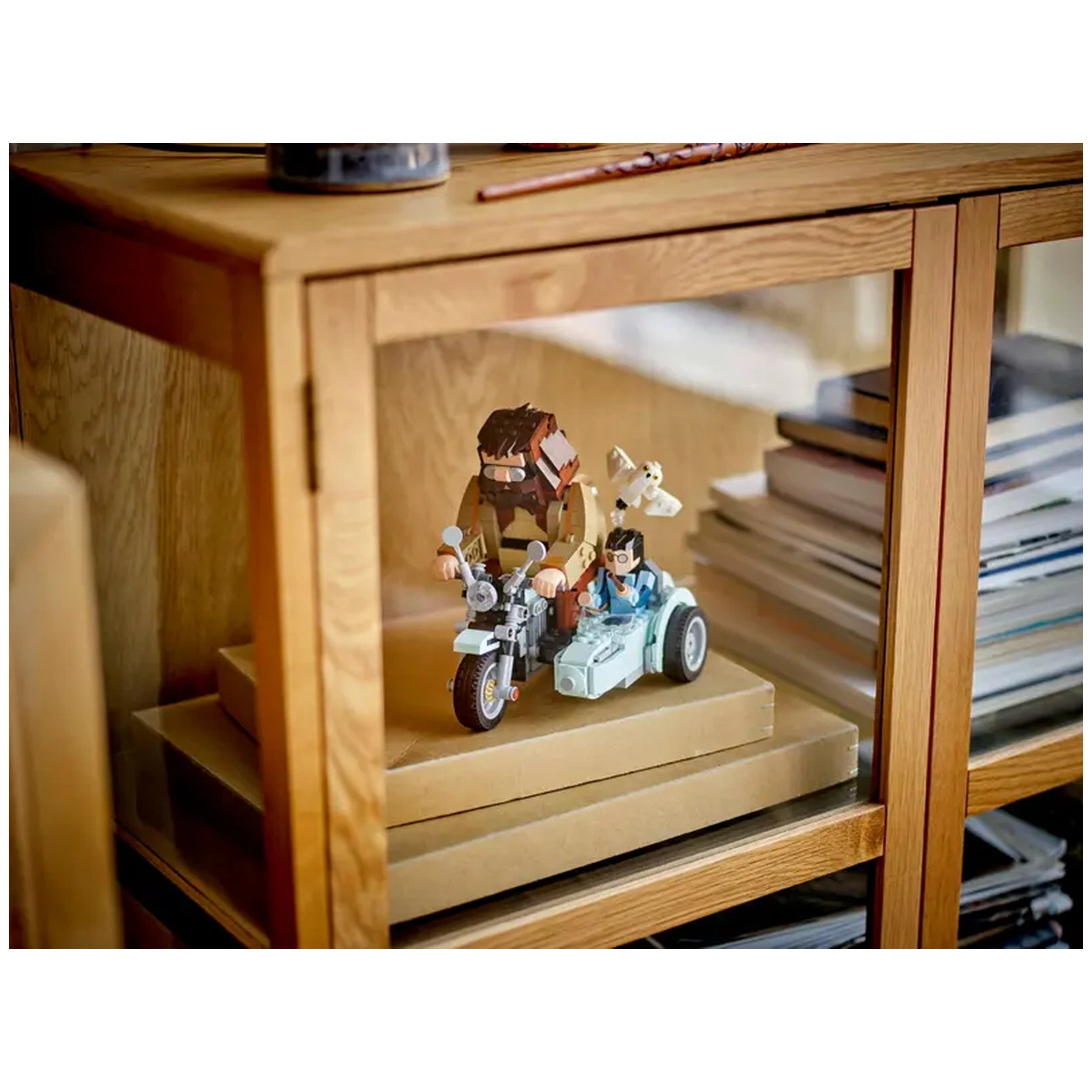 LEGO set features Hagrid and Harry on a blue motorcycle with a sidecar, Hedwig flying above, displayed in a cozy wooden cabinet.