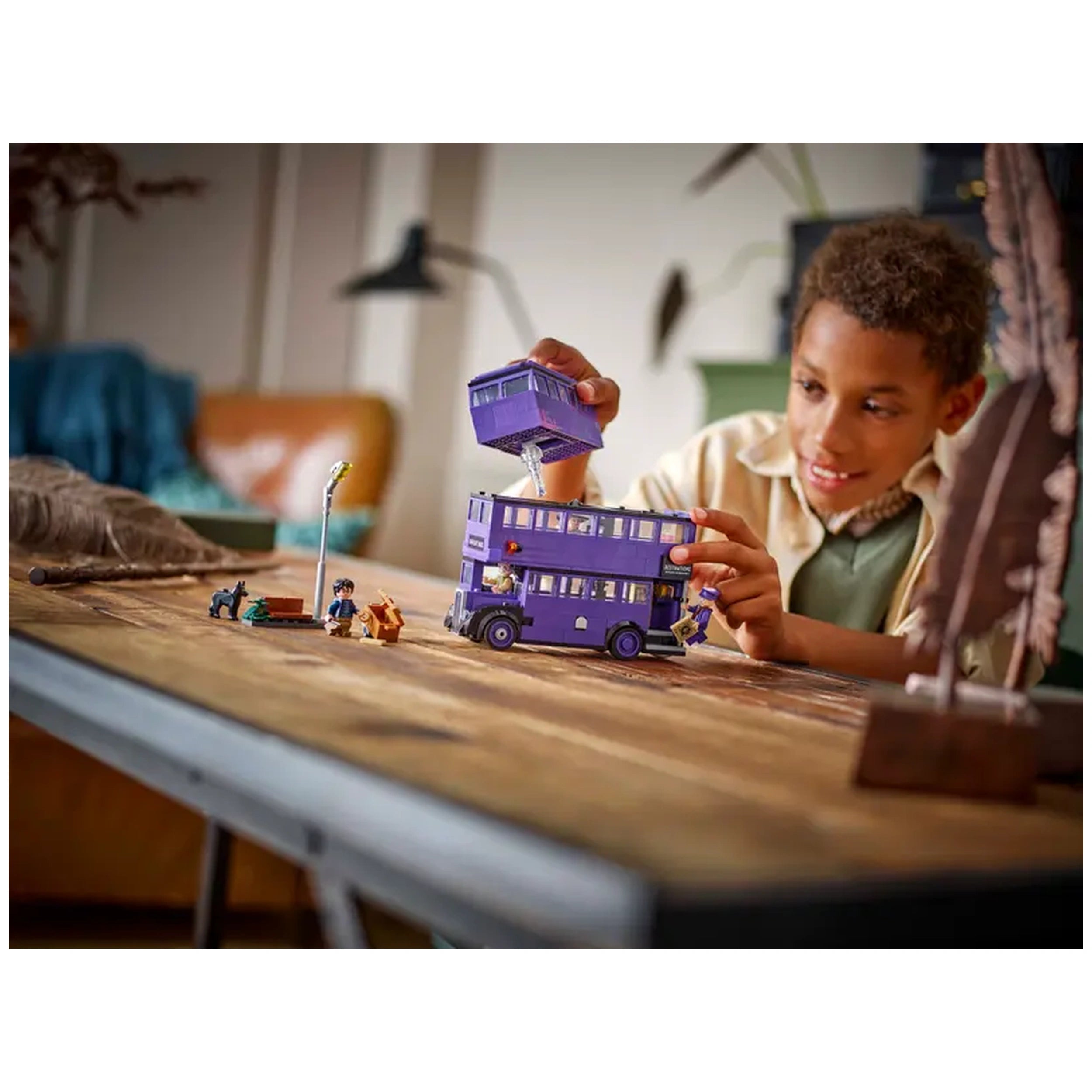 A child joyfully interacts with the purple LEGO® Harry Potter™ Knight Bus™ construction set, lifting the upper deck to reveal detailed interior play features on a wooden table.