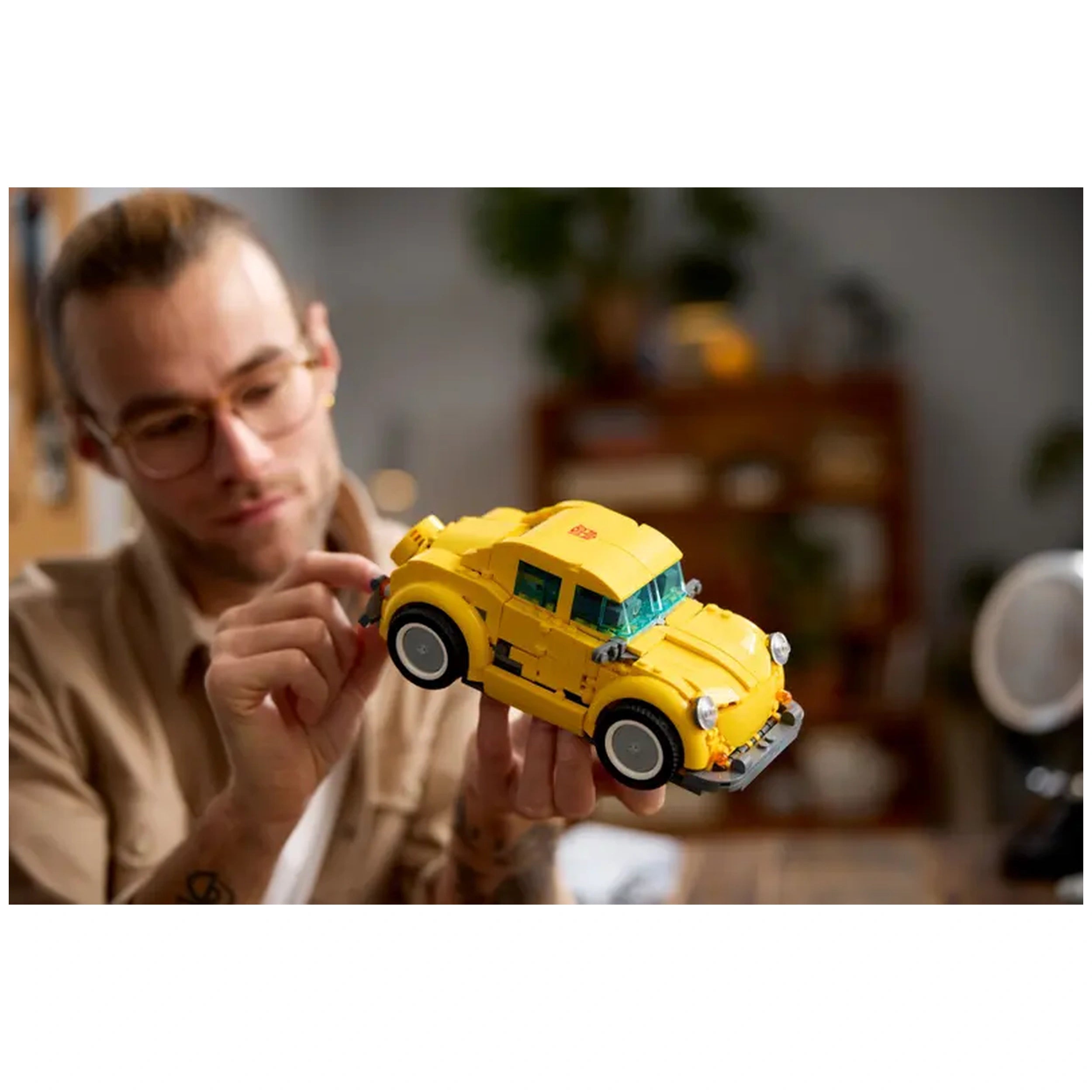 Adult male with glasses inspecting a yellow LEGO® Icons Bumblebee construction set, resembling a vintage car, in a cozy indoor setting.