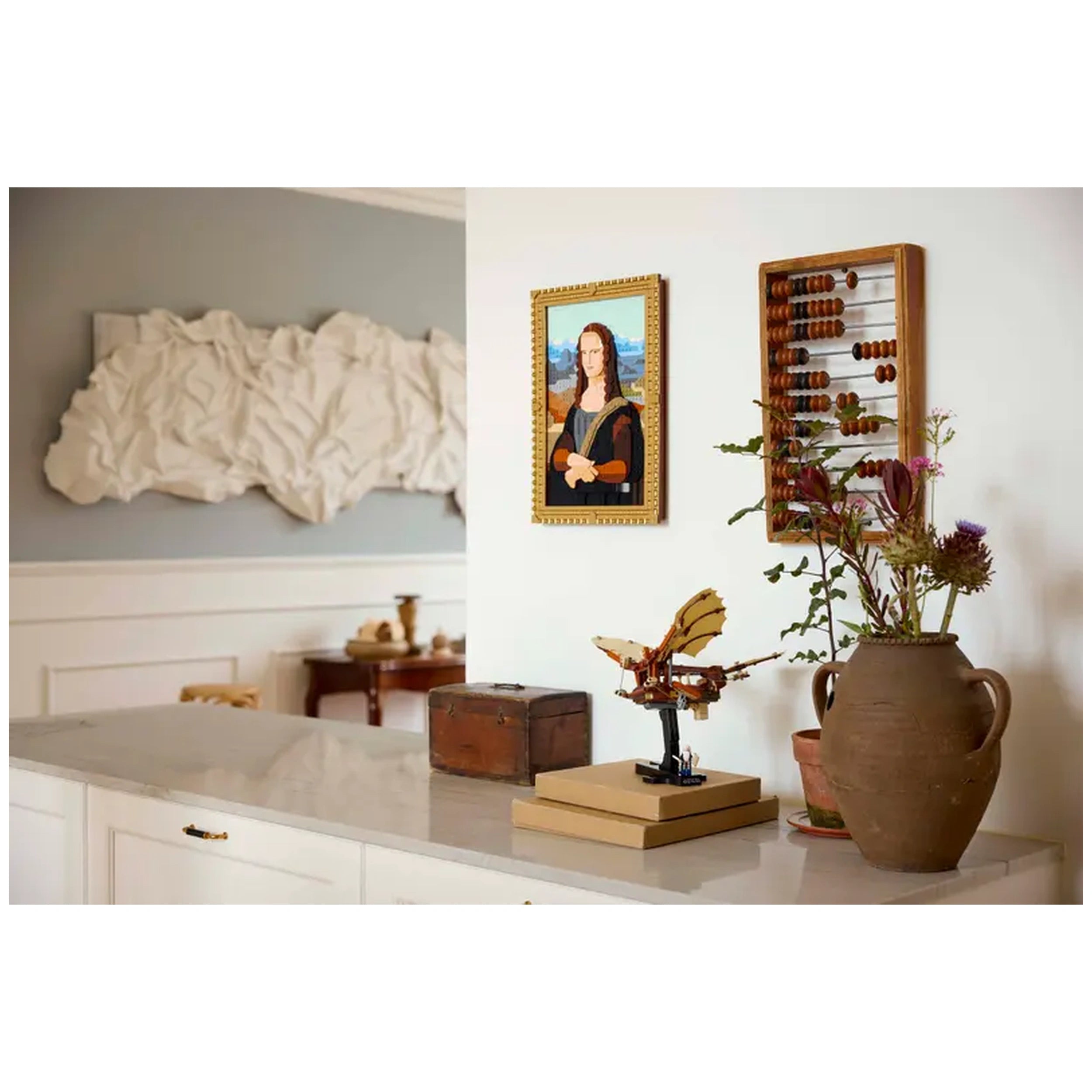 LEGO® Icons Leonardo da Vinci's Flying Machine displayed on a marble surface beside a vintage wooden box and potted flowers. A framed portrait, inspired by the Mona Lisa, hangs on the wall, next to an abacus.