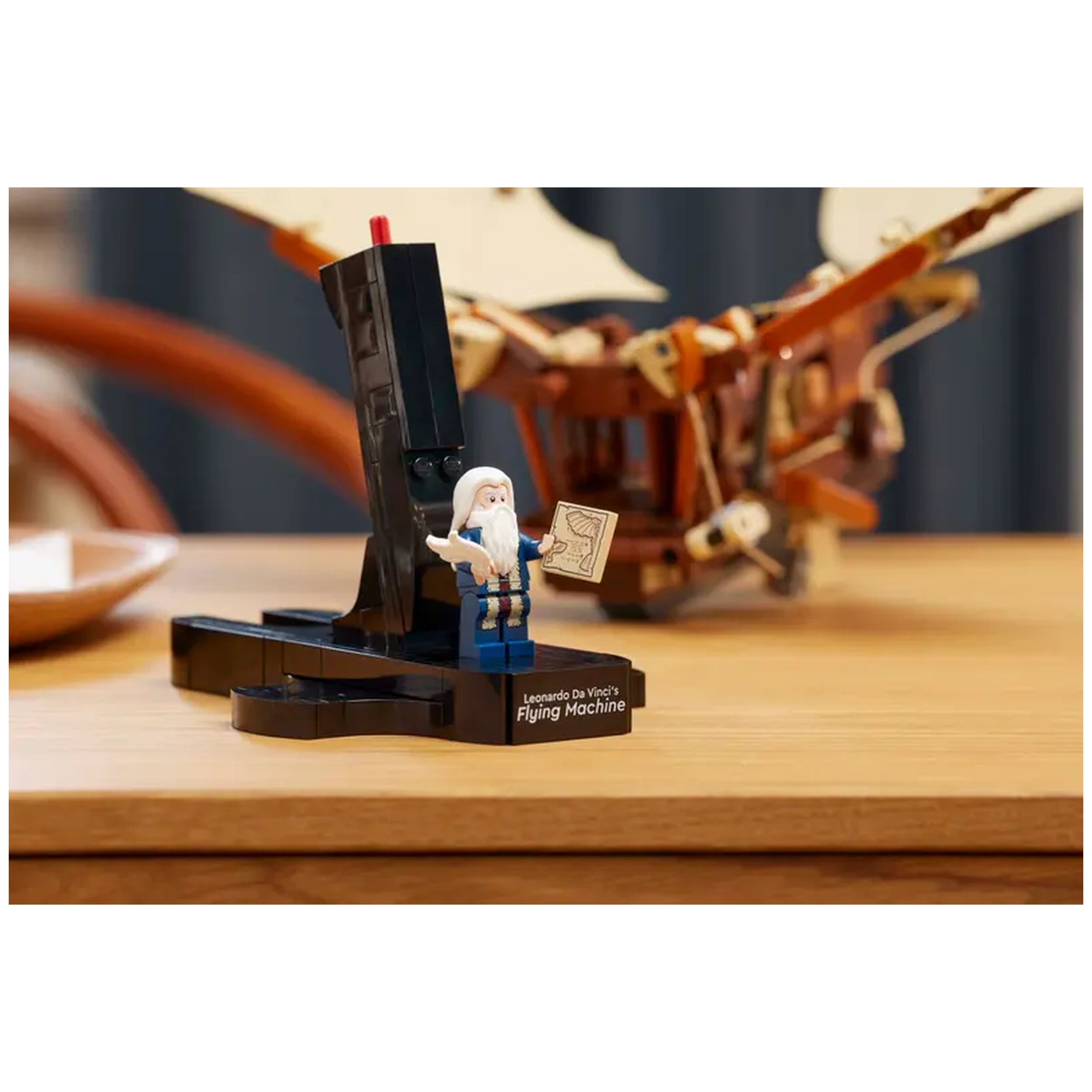LEGO® Icons Leonardo da Vinci's Flying Machine set features a detailed model with a flying machine backdrop, a mini figure of Da Vinci holding a drawing, and a sturdy black base on a wooden table.