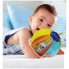 Baby smiles while holding a colorful plush toy on a white bed with a blue pillow, wearing a plaid shirt.