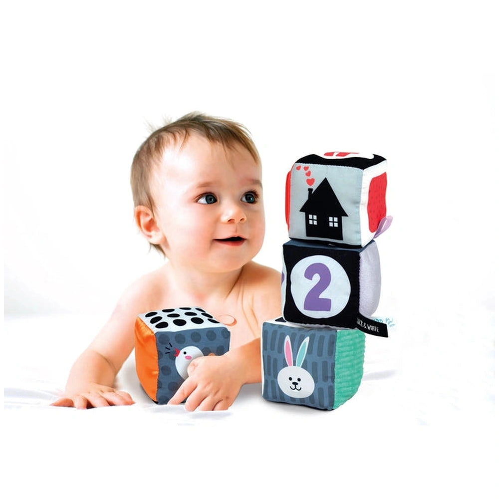 Baby smiling while sitting on a white surface, reaching for colorful soft activity blocks with various patterns.