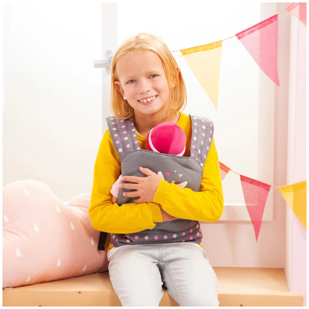 BAYER DESIGN Doll Carrier in gray and pink, child in yellow shirt holding a stuffed animal, sitting on a wooden bench.