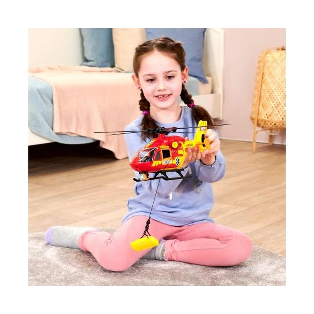 Dickie ambulance helicopter toy in girl's hands, featuring red and yellow colors, set in a cozy bedroom with pink accents.