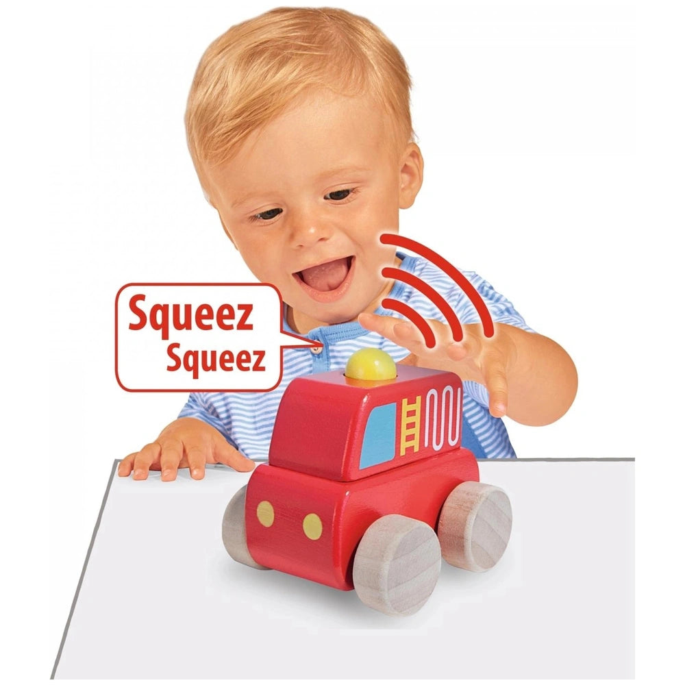 A smiling child with blonde hair holds a red toy truck with a yellow top, sitting at a table with a white surface.