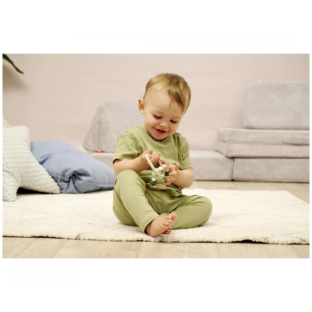 Eichhorn Baby HiPP Grapper is held by a smiling child in a green outfit, sitting on a white rug with a blue pillow nearby.