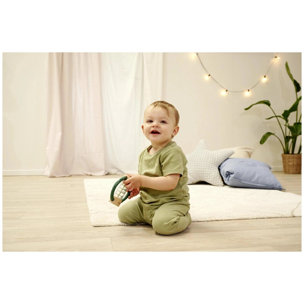 Eichhorn Baby HiPP Sound Rattle in a babys hand, set in a bright nursery with a white rug and wooden floor.