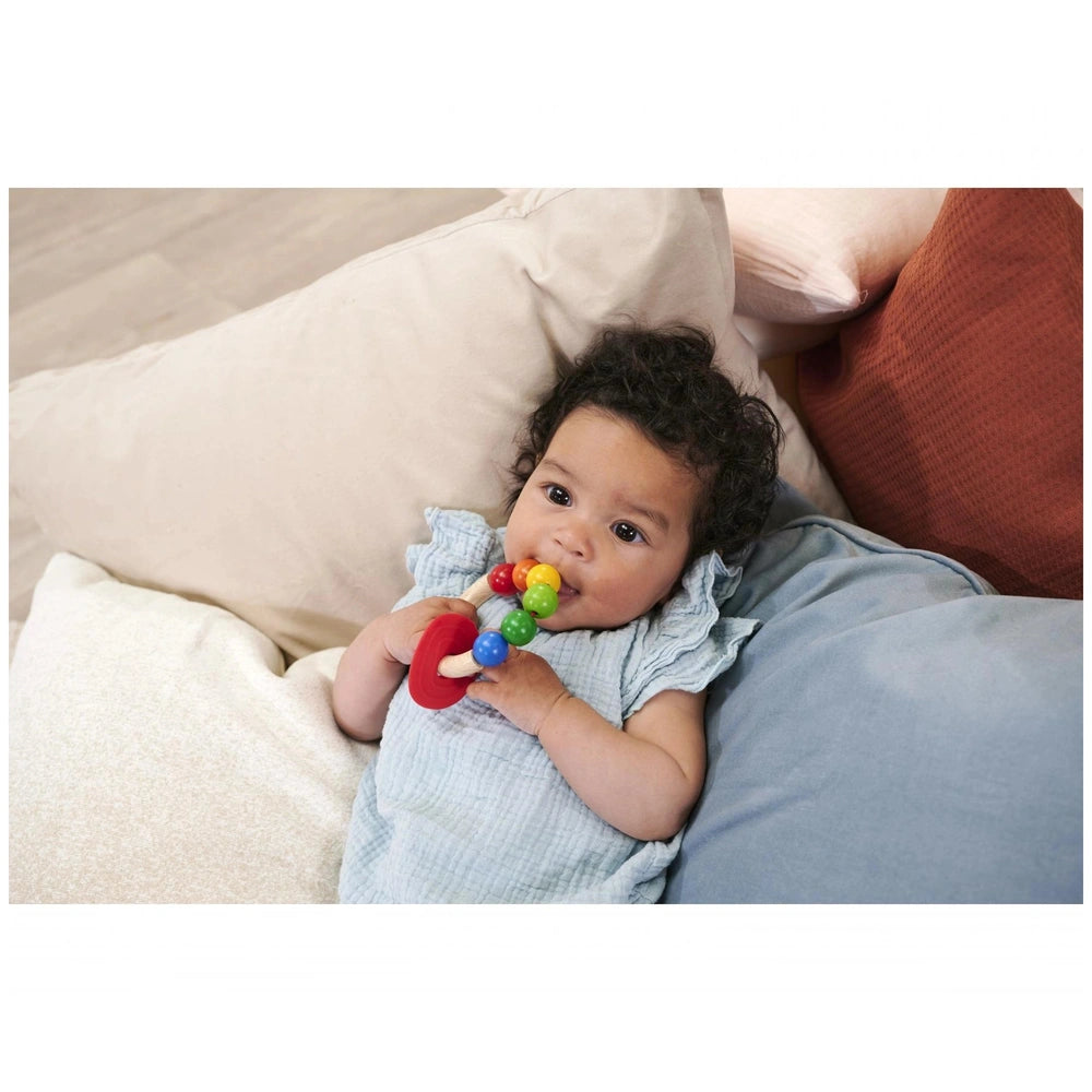 Eichhorn Eh Baby Wooden Teething Ring held by a baby on a couch, showcasing a colorful design and a warm background.