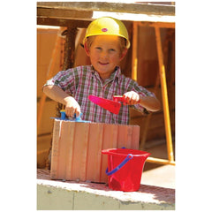 Child in a yellow hard hat holds a red shovel and blue trowel, standing by pink bricks and a yellow ladder.