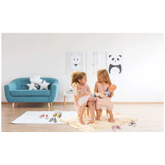 Heless pink sneakers for dolls are showcased in a lifestyle scene with two girls, a blue couch, and a light rug.