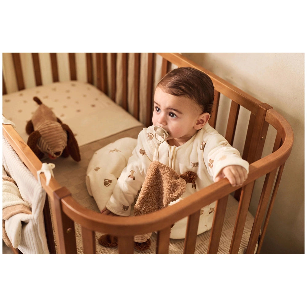 Jollein pacifier cloth features a baby in a crib, surrounded by a brown blanket and stuffed animal on a patterned white mattr