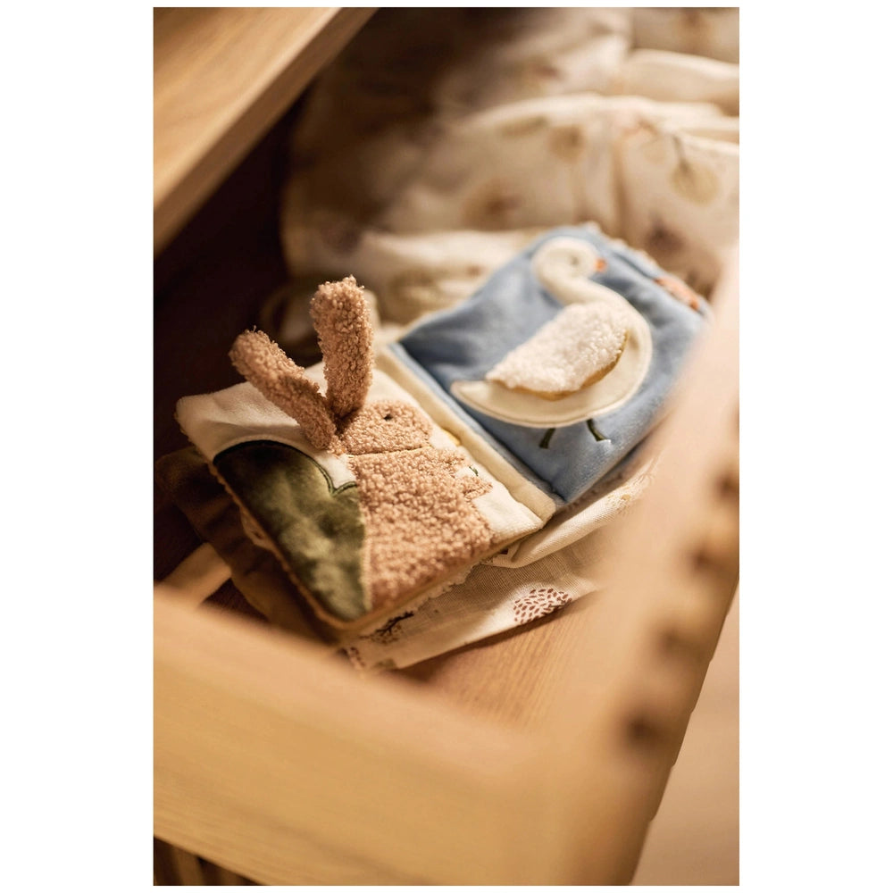 Jollein soft activity book featuring a blue patterned cloth, brown stuffed animal, and decorative paper on a wooden surface.