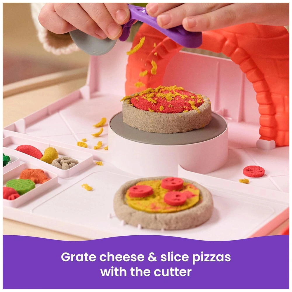 Kinetic Sand pizza playset features a child using a purple spatula to spread cheese on a colorful pizza in a play kitchen.