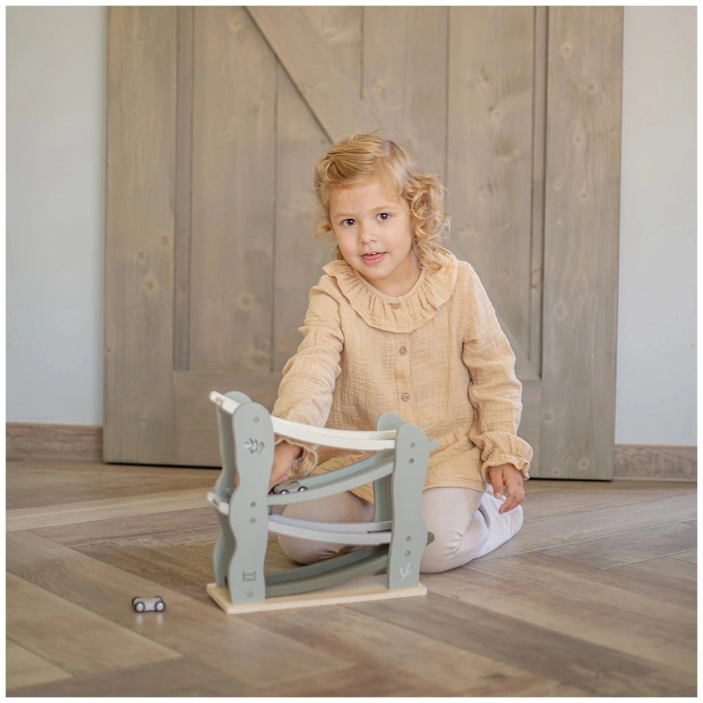 Blonde child in a beige dress plays with a light blue Label-Label car slider toy on brown wooden flooring.