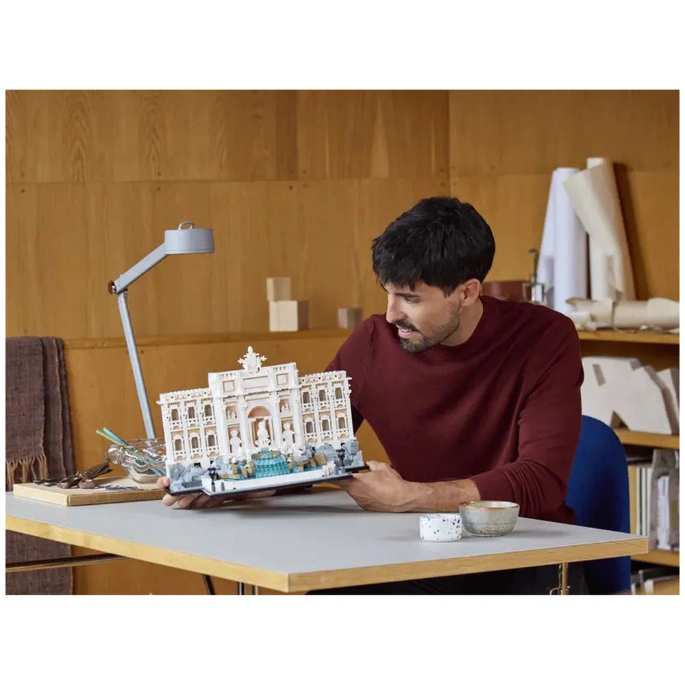 Lego Architecture Trevi Fountain model is held by a focused man seated at a light wooden table in a workshop setting.