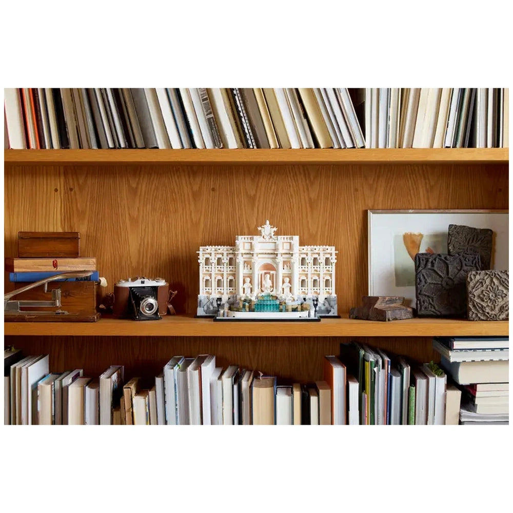 Lego Trevi Fountain construction set displayed on a wooden bookshelf alongside colorful books and decorative items.