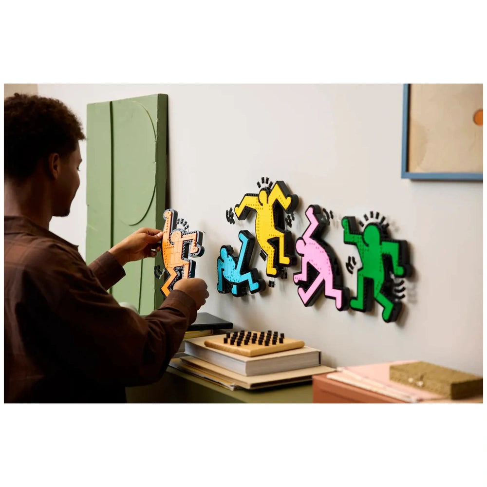 LEGO Art kit featuring colorful cutouts of Keith Haring figures in motion on a white wall, with a person holding a small obje