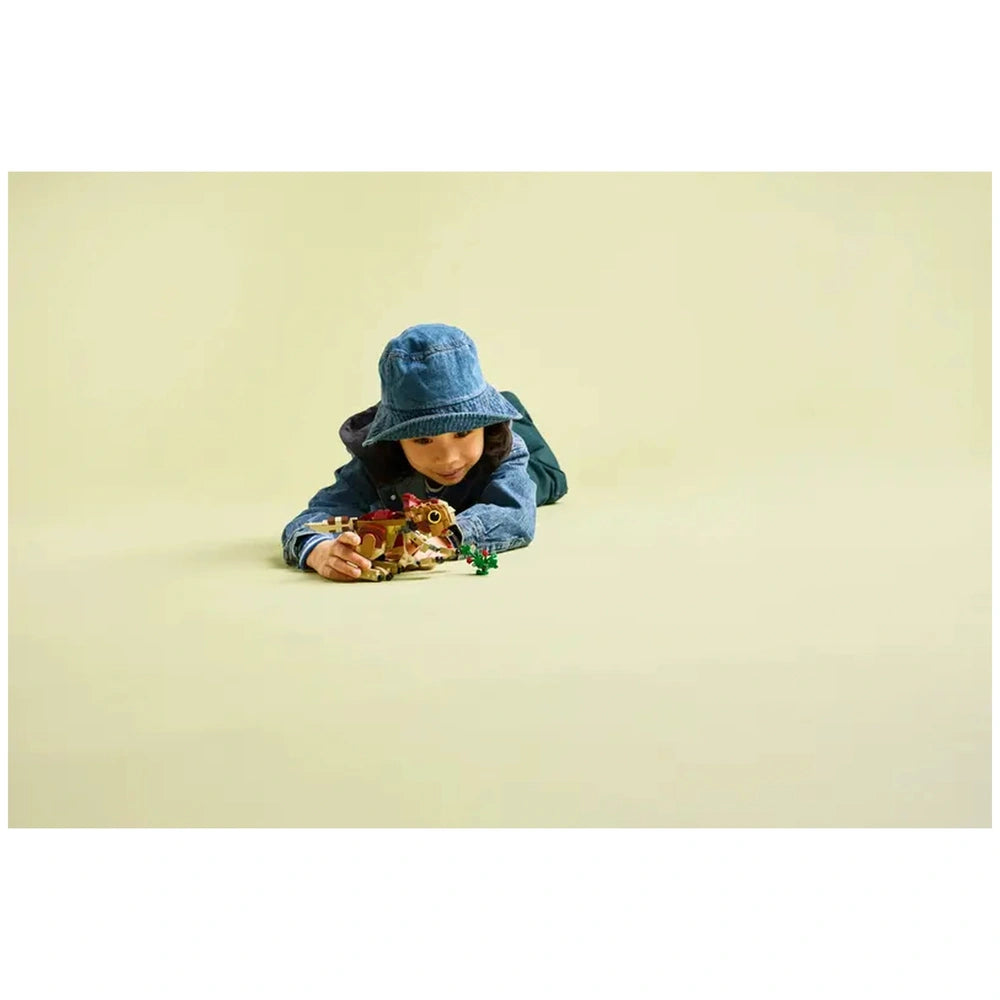 A child wearing a denim jacket and blue bucket hat lies on a light yellow background, playing with the LEGO Baby Dinosaur Dolores set and colorful plant pieces.