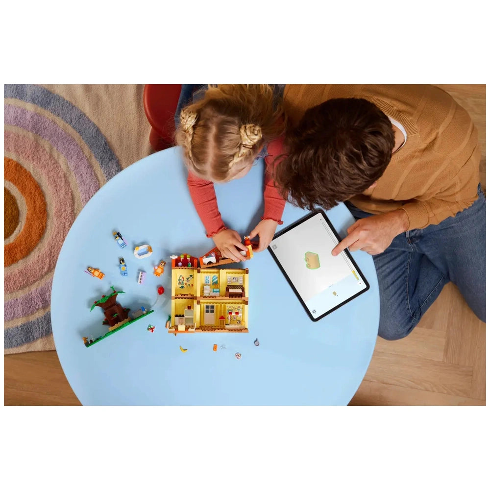 A young child and an adult are engaged in play at a light blue table, focusing on the LEGO Bluey’s Family House set. The child is assembling characters and accessories, while the adult uses a tablet for guidance. Surrounding them are scattered playset pieces and a colorful rug underneath.