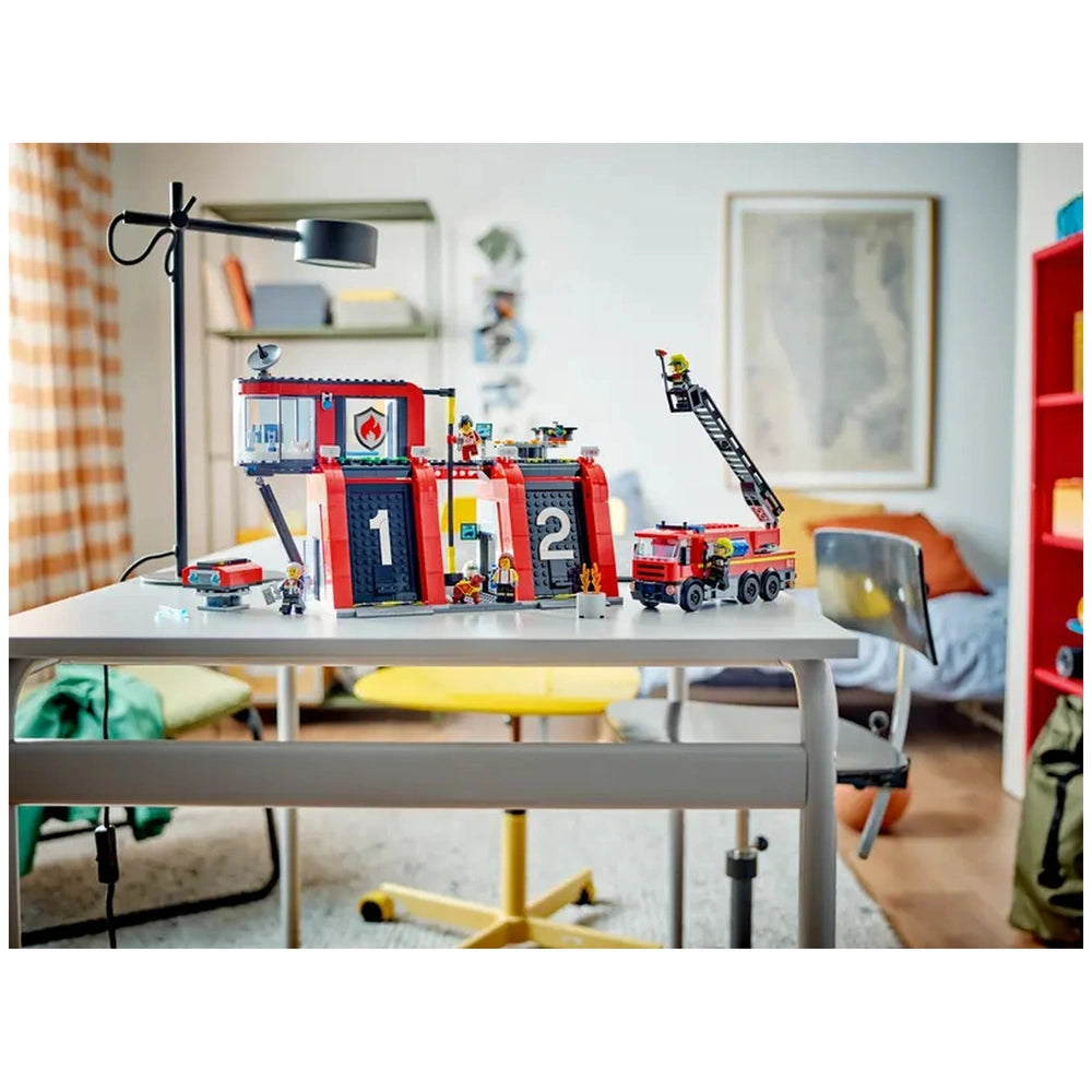 LEGO® City Fire Station set on a table, featuring a red building with numbered garage doors, a fire truck with a ladder, and five firefighter minifigures. A lamp and colorful furniture are visible in a cozy room.