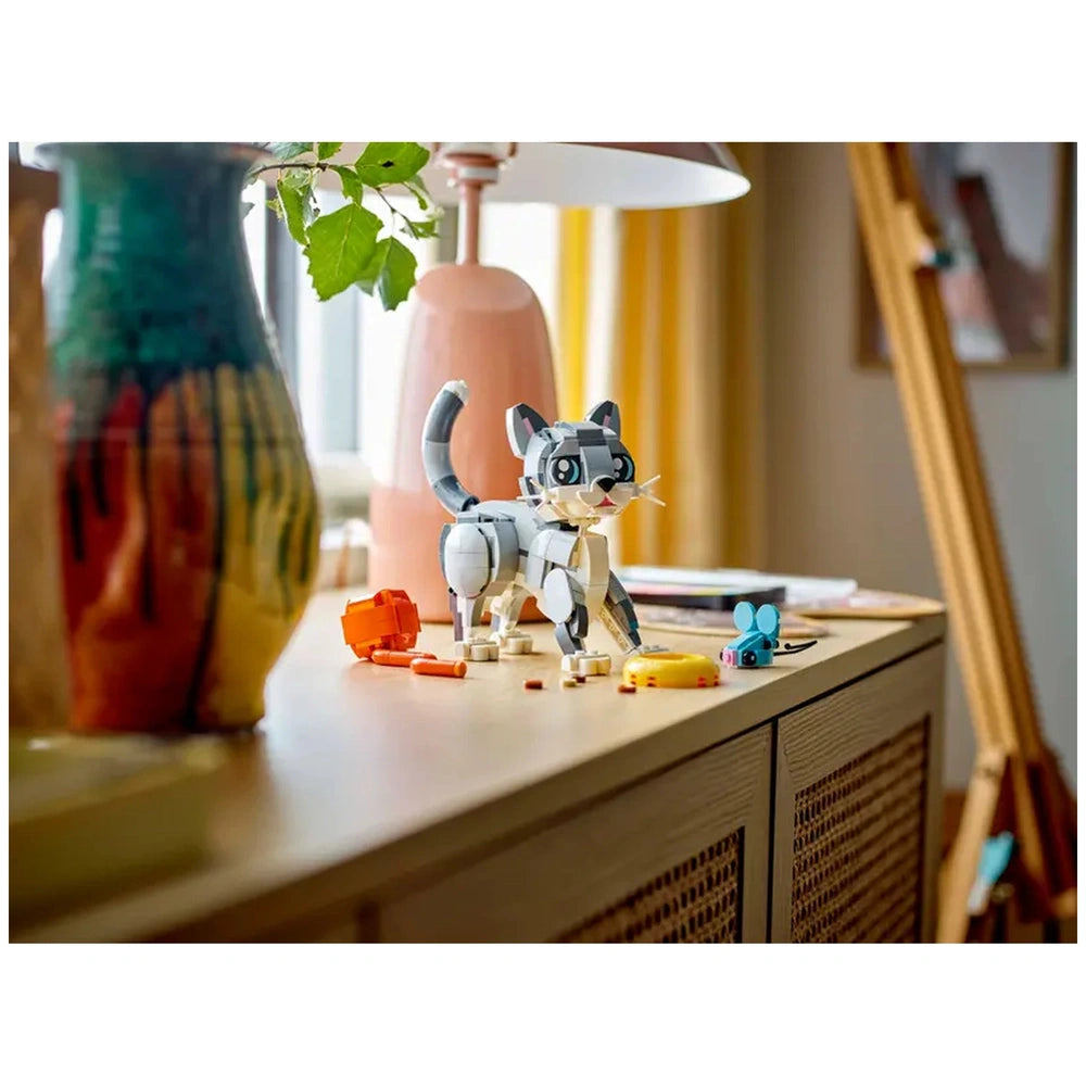LEGO Creator 3in1 Playful Cat set displayed on a wooden table, featuring a gray cat toy, toy mouse, food bowl, and yarn ball, surrounded by colorful decorations.