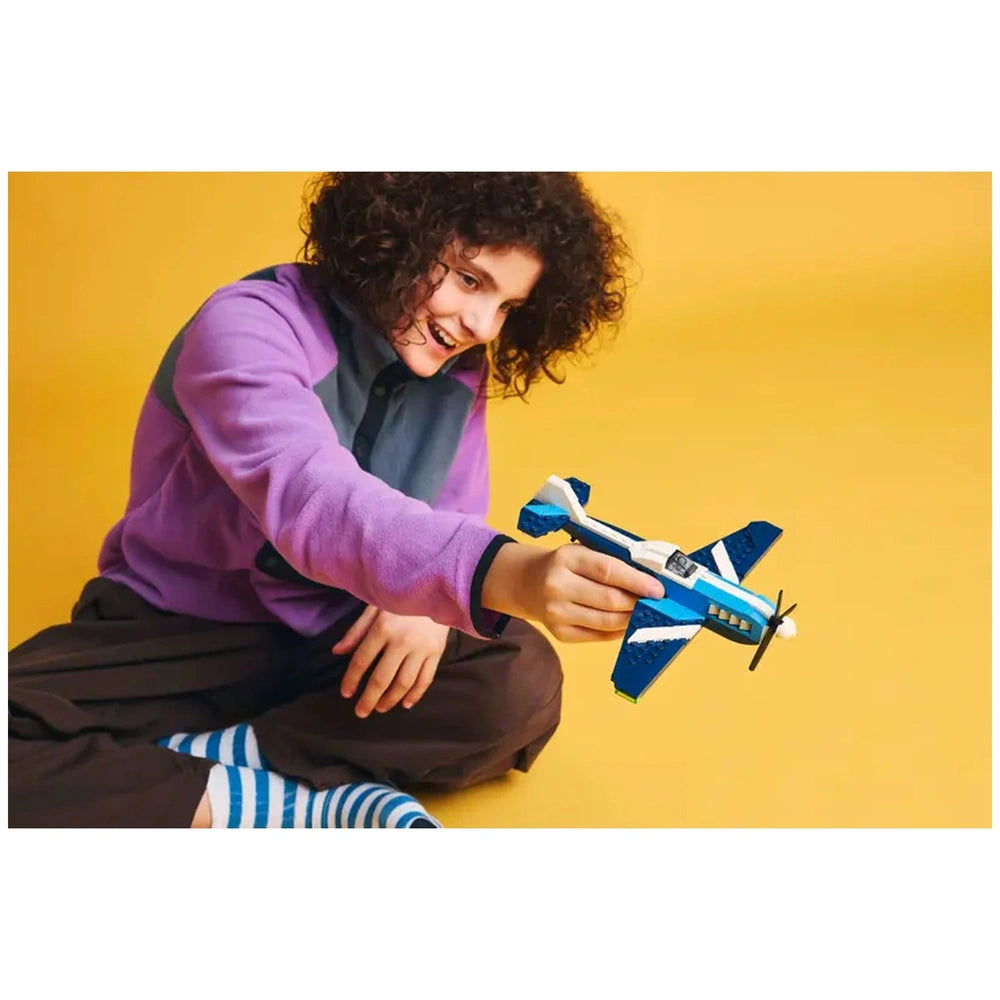 A child with curly hair, wearing a purple and gray sweatshirt, smiles while holding a blue and white LEGO® race plane in a playful pose against a bright yellow background.