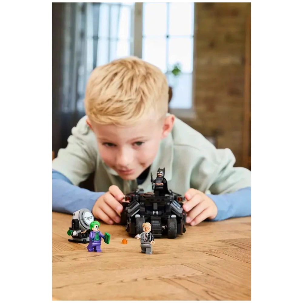 A child focuses intently on a LEGO Batman Tumbler set, featuring the Batmobile and minifigures of the Joker and Harvey Dent.