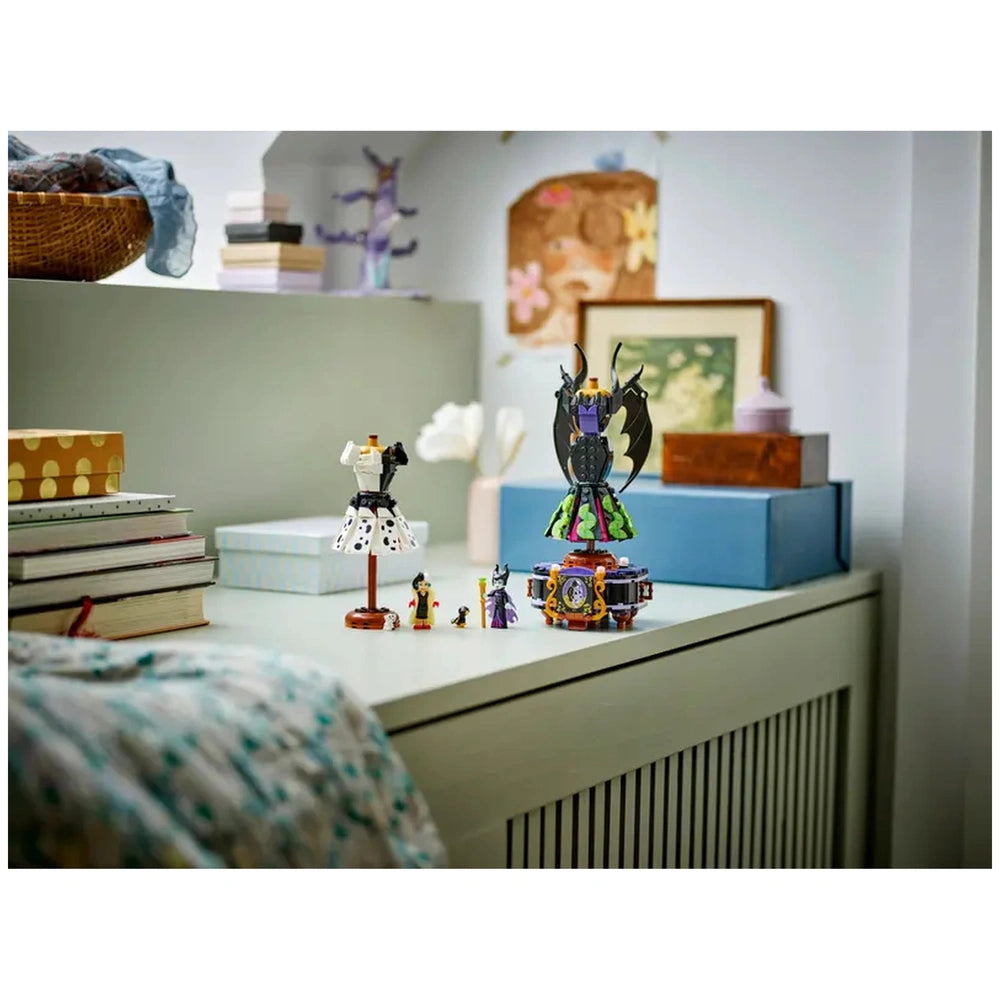 Two buildable dresses representing Maleficent and Cruella De Vil displayed on a dresser, surrounded by LEGO minidolls and accessories, with books and decorative items in the background.